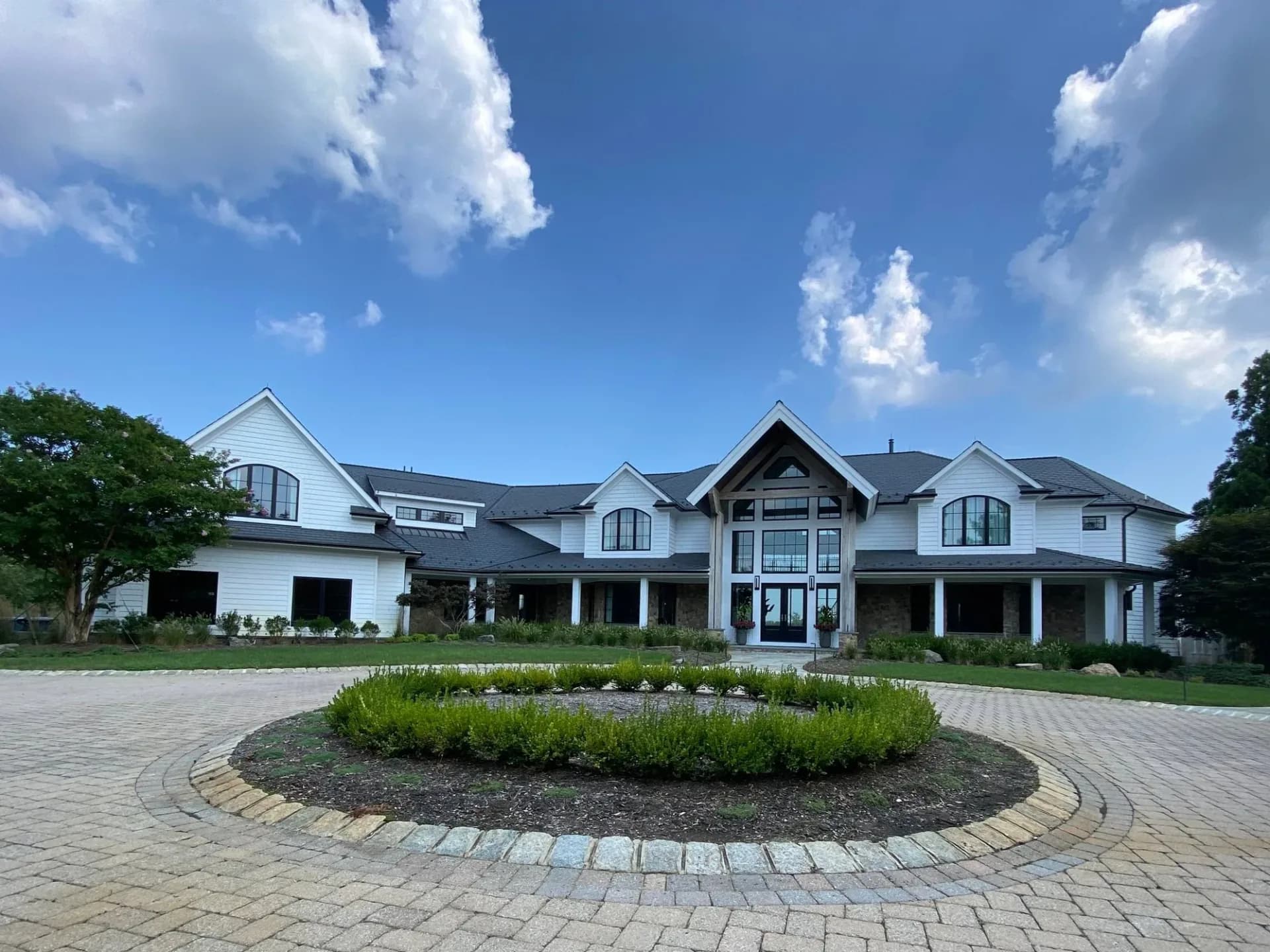 Modern farmhouse exterior with white and dark facade, circular paver driveway with landscaping feature, and attached garage by Shayna Vrabel.
