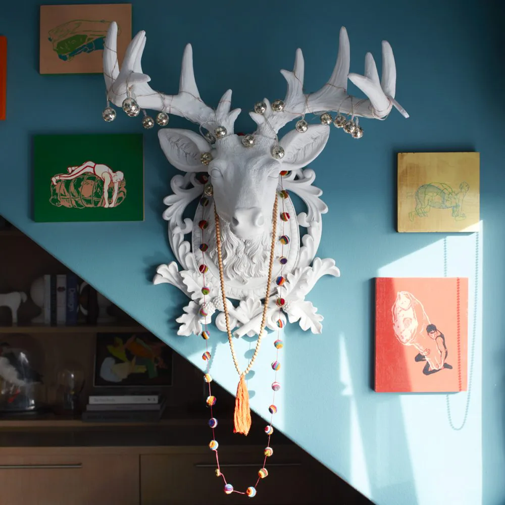 Turquoise bedroom wall featuring a white decorative deer head with ornamental details and hanging jewelry, surrounded by framed artwork by Sheila Bridges.