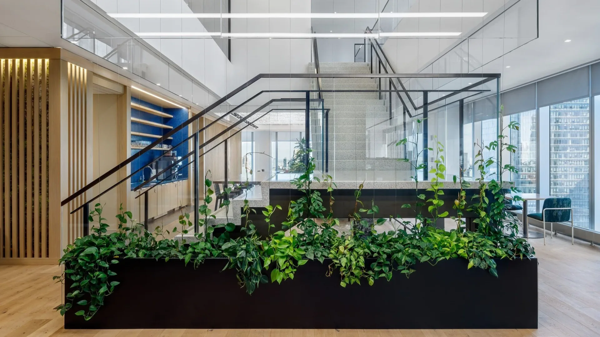 Modern office atrium with floor-to-ceiling glass partitions, wooden slatted accent wall, cascading greenery in black planter boxes, and natural light by Shereese Trumpet.