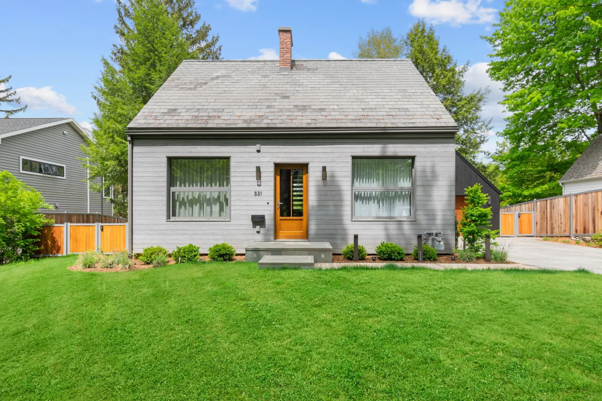 A renovated mid-century cottage with gray siding, yellow door, and symmetrical windows sits on manicured lawn with mature trees by Shereese Trumpet.