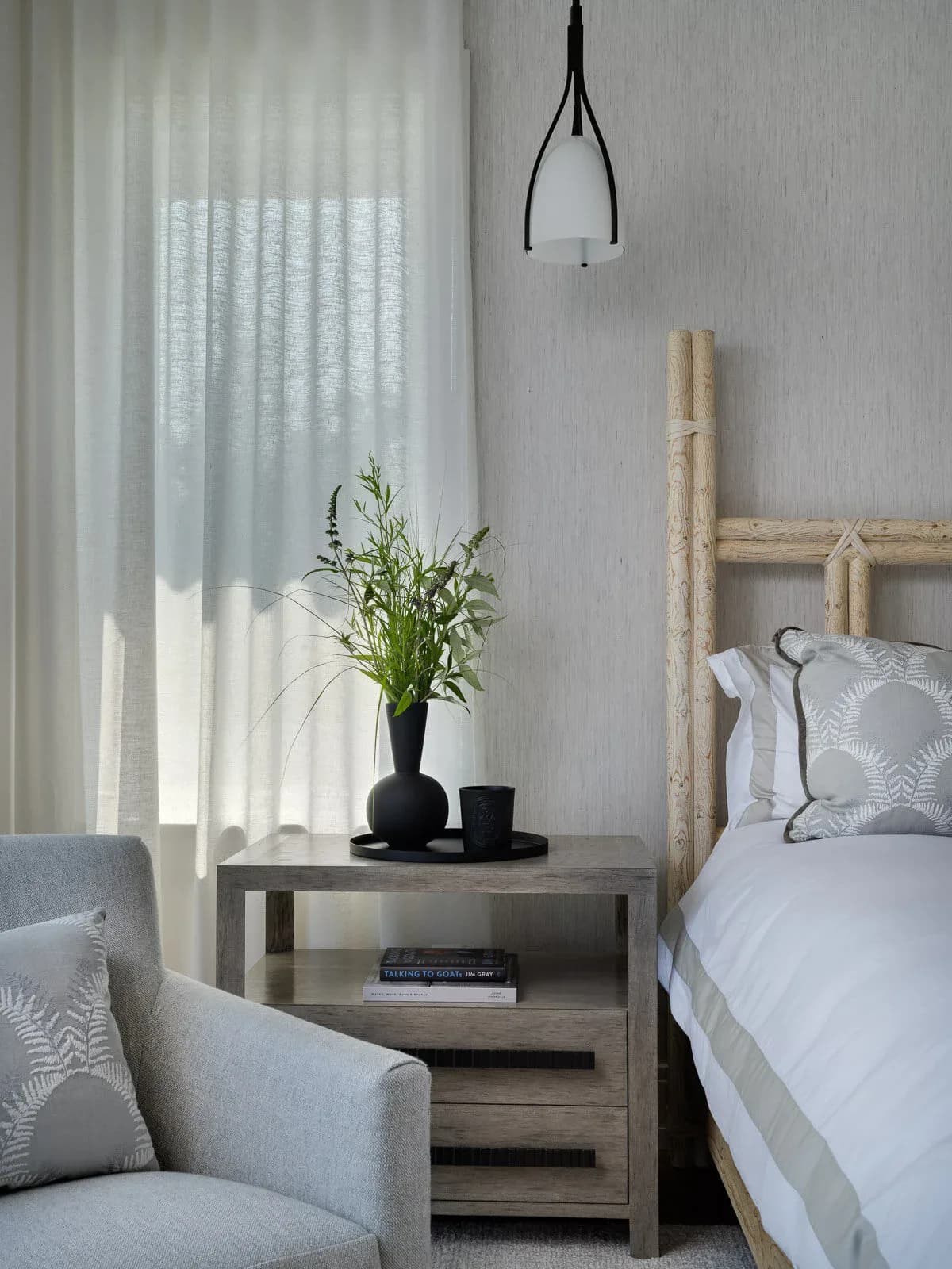 Serene bedroom corner featuring a natural wood bed frame, gray upholstered chair, wooden nightstand with black vase, pendant lighting, and sheer curtains by Sherry Brous.