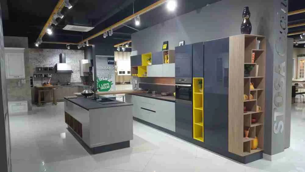 Modern kitchen showroom with white cabinetry, dark upper units, yellow accent storage, central island, and polished concrete flooring by Bassel Shihab.