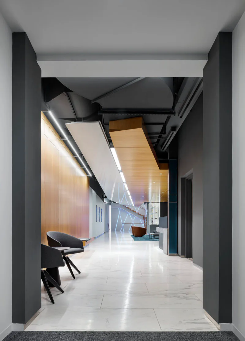Modern corridor with gray concrete walls, polished white floor, and warm wooden ceiling panels, featuring contemporary black furniture and linear lighting by Shurouq Al Jammal.