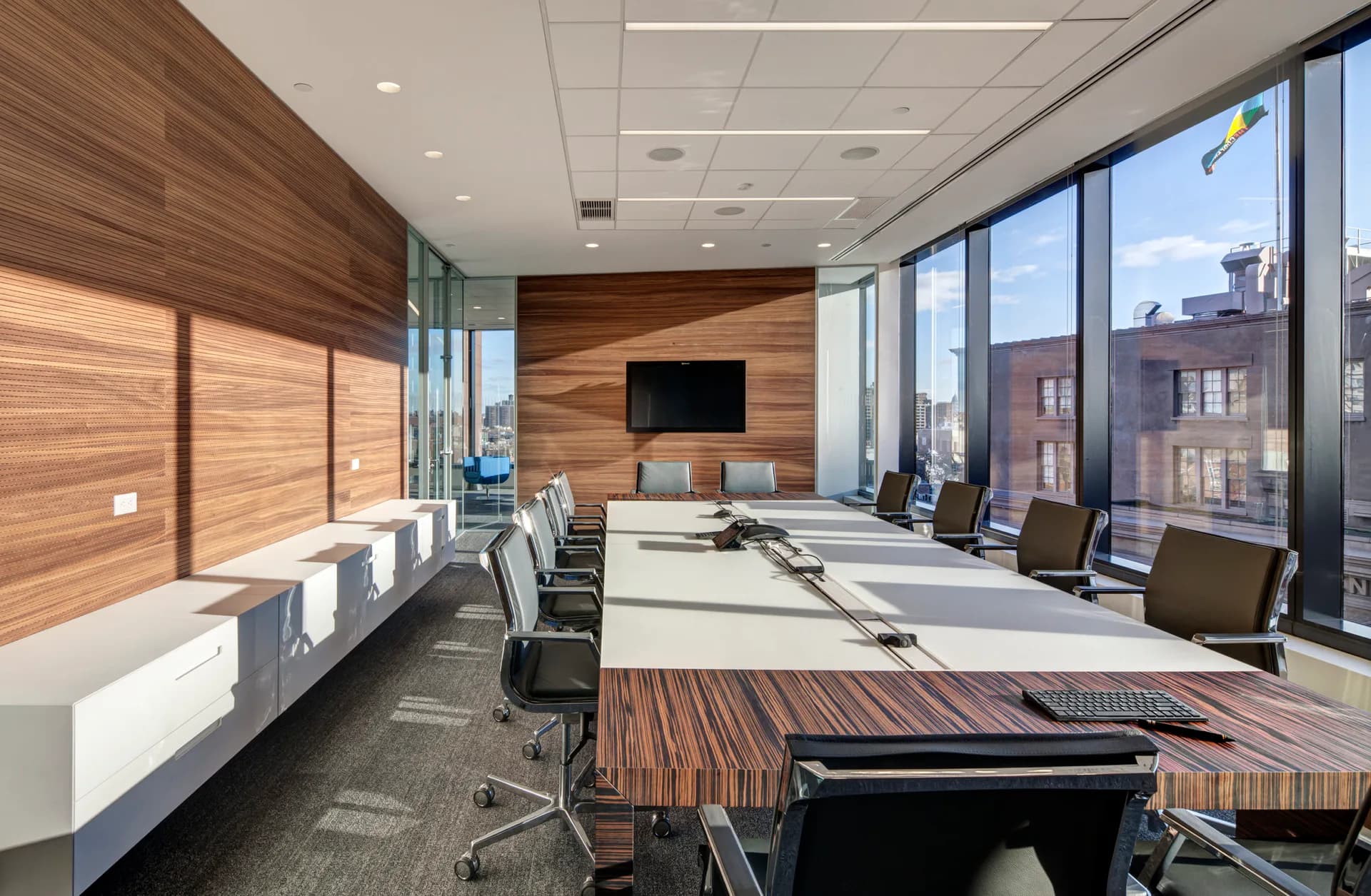 Modern office space with wood-paneled accent wall, white built-in shelving, long work table, and floor-to-ceiling windows overlooking city by Shurouq Al Jammal.