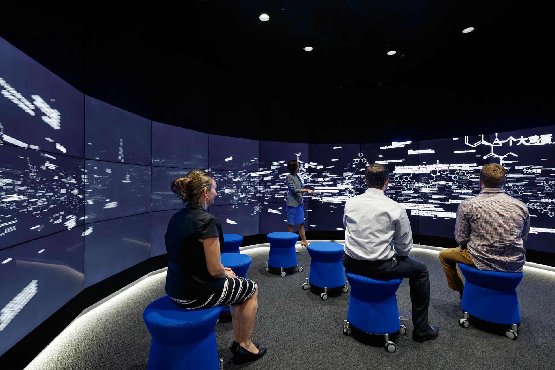 Immersive exhibition space with curved walls displaying illuminated aerial cityscape imagery, blue seating elements, and ambient lighting design by Shurouq Al Jammal.