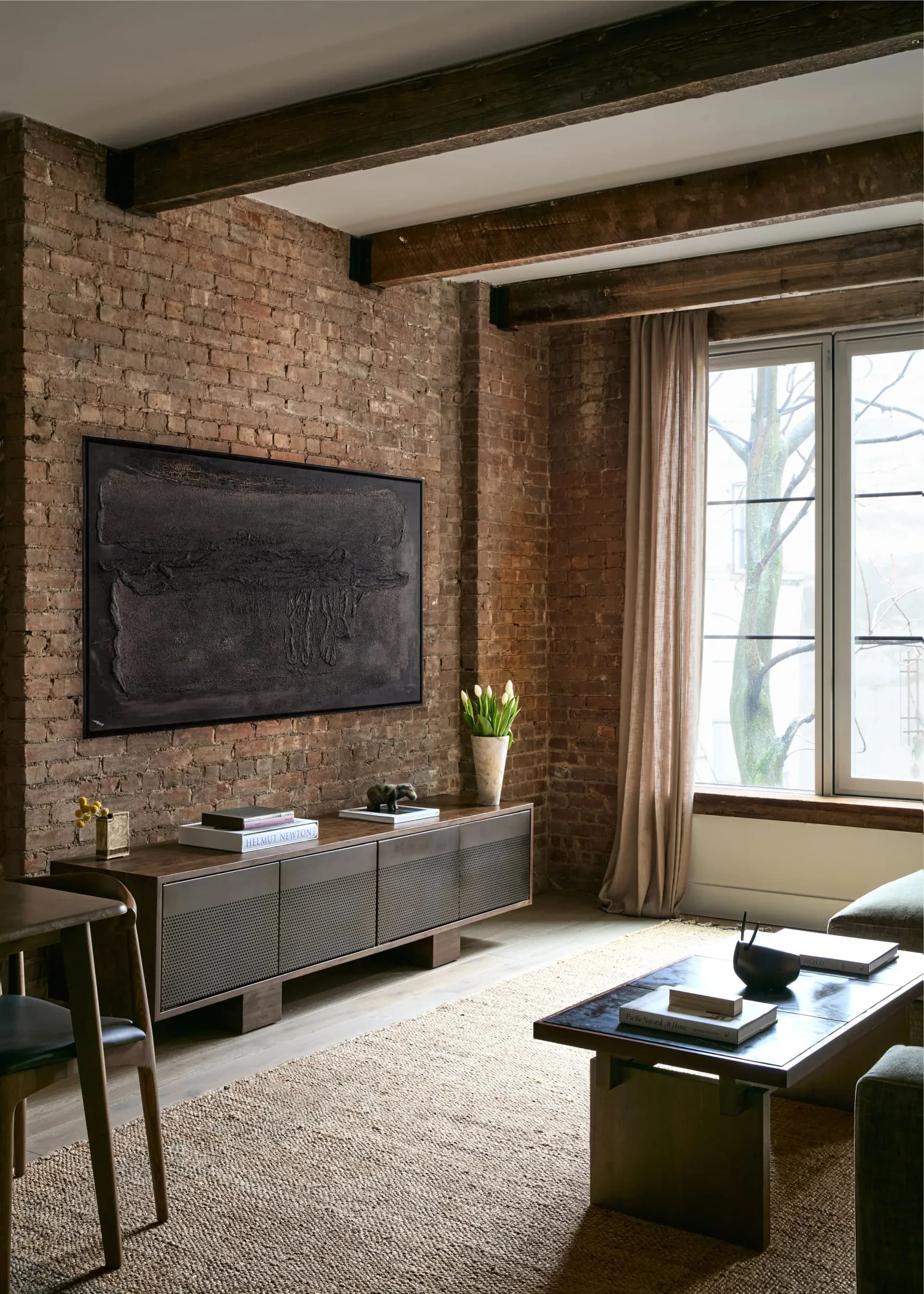 Modern loft living room with exposed brick walls, dark wood beams, mounted TV, contemporary credenza, and large windows with neutral curtains by Simone Lawton.