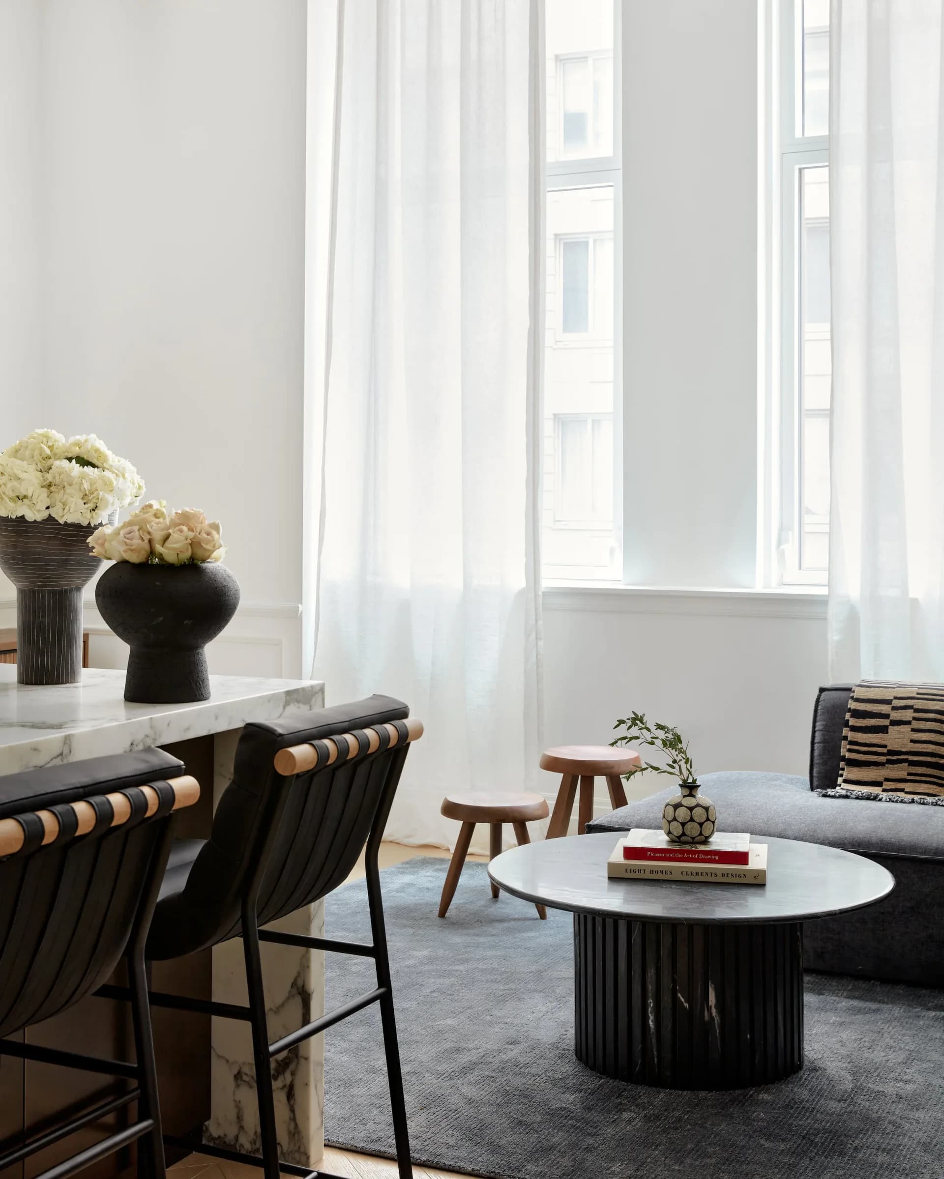 Bright modern living room with minimalist black and white palette, featuring black sculptural furniture, round white coffee table, and flowing white curtains framing tall windows by Simone Lawton.