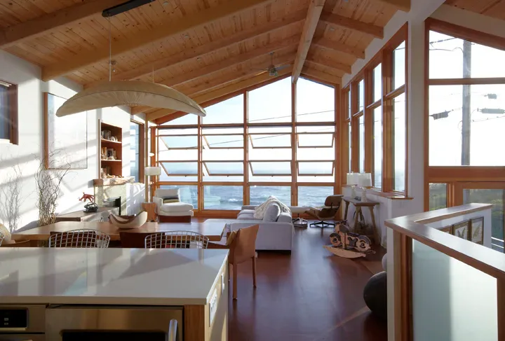 Bright modern interior with exposed wood ceiling beams, extensive glazing overlooking water views, and warm wood flooring by Sisa Suriel.