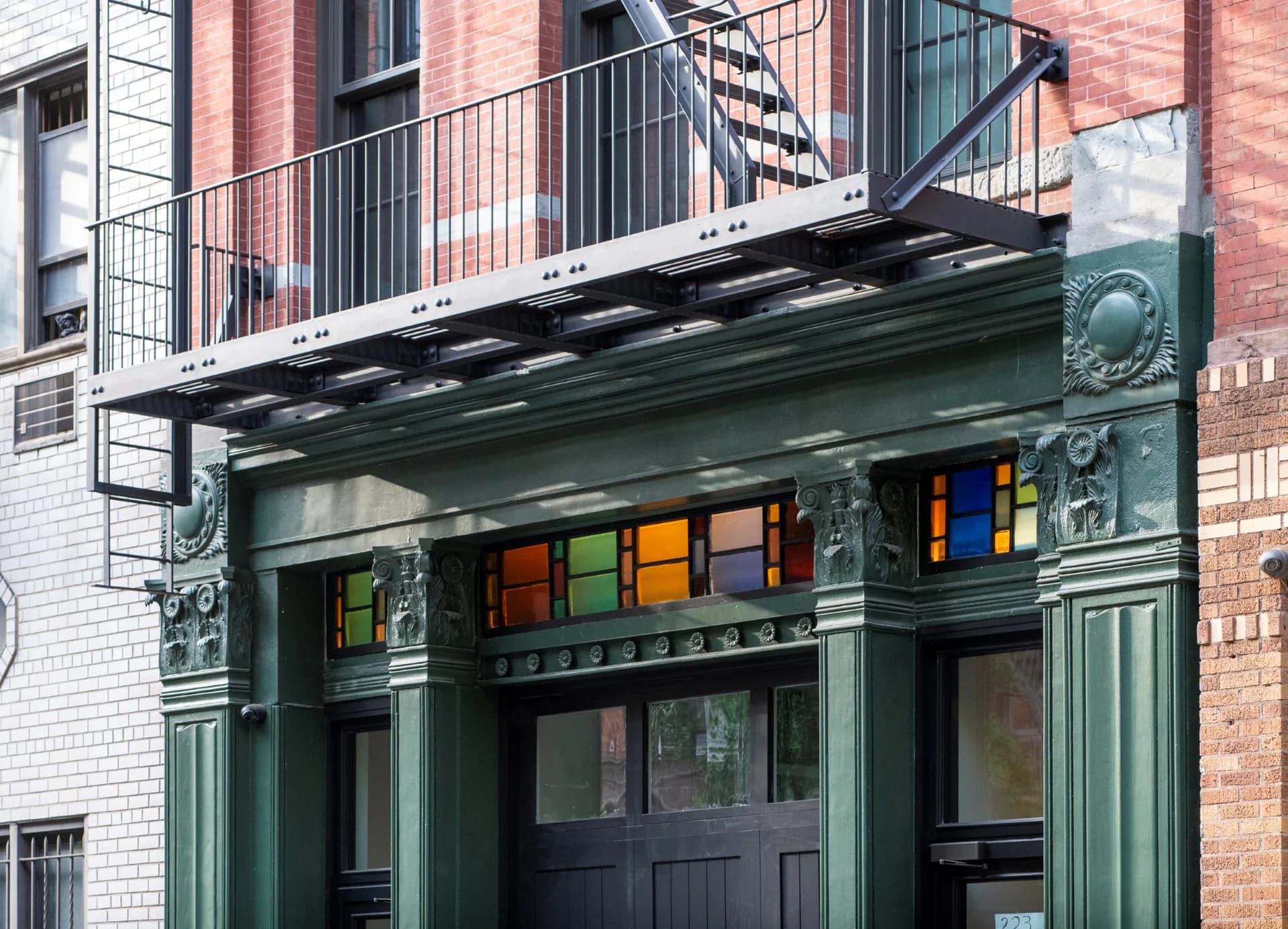 Historic storefront with green ornamental metalwork facade and colorful transom windows above dark entrance doors, designed by Sohui Kim.