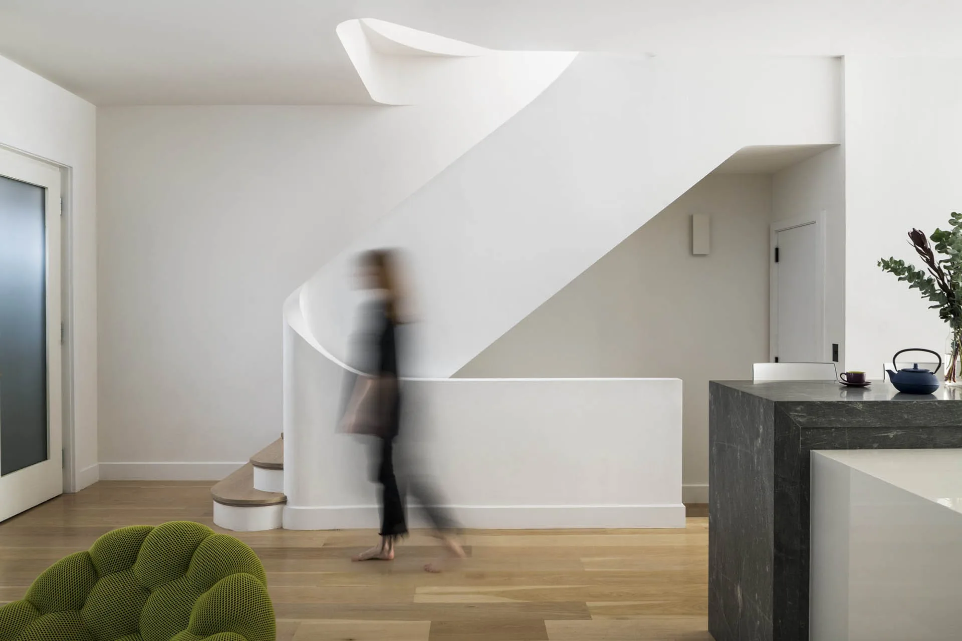 Modern spiral staircase with white walls and wooden flooring, featuring a blurred figure in motion, minimalist interior design by Sohui Kim.