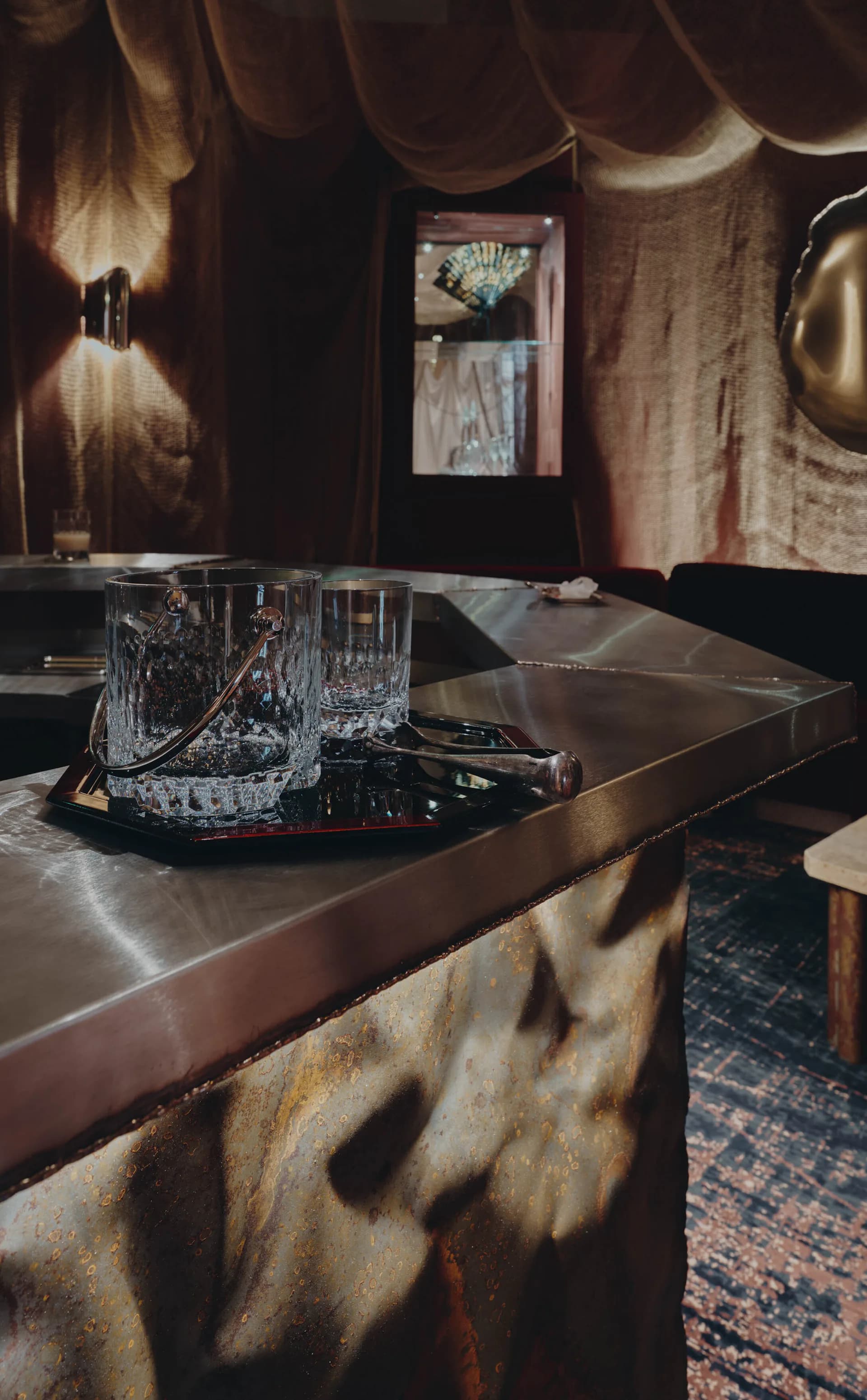 Warm-toned interior with brass table, crystal glassware, draped textiles, and layered lighting creating an intimate hospitality space by Sophie Dries.