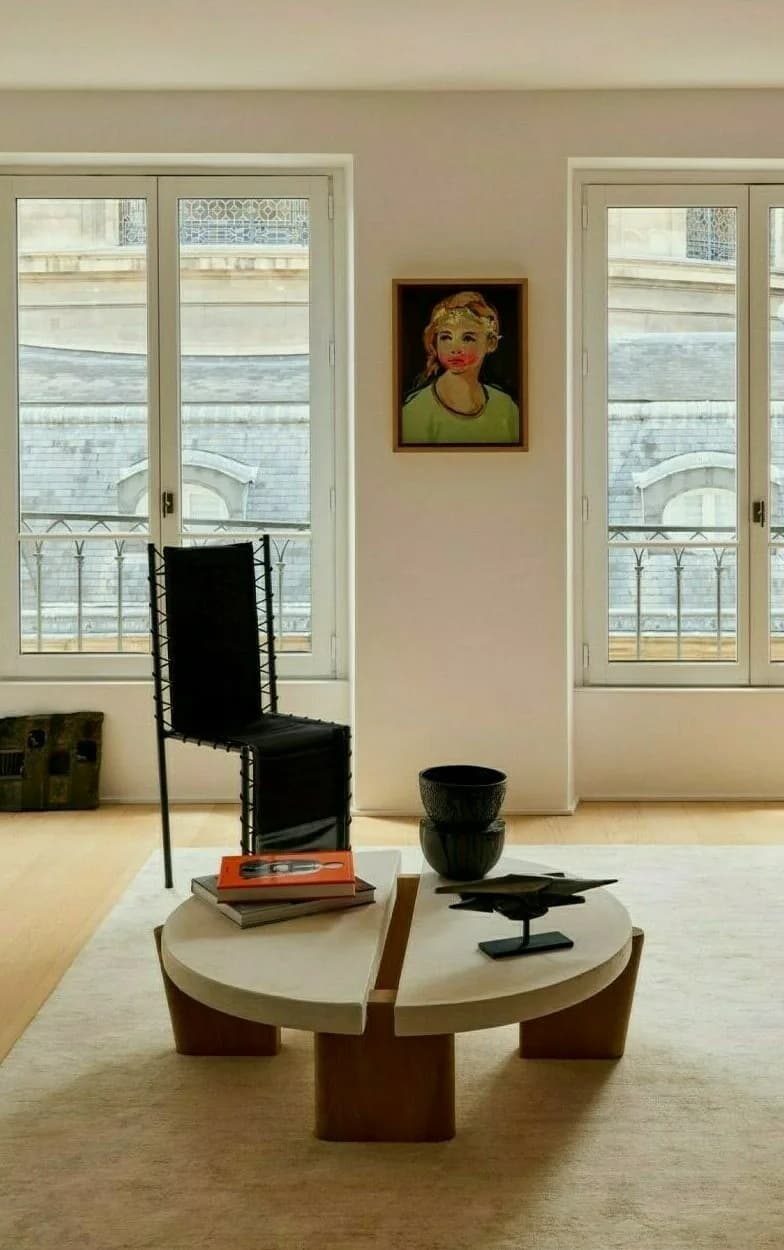Minimalist interior with round wooden table, black chair, and French doors opening to Parisian balconies, designed by Sophie Dries.