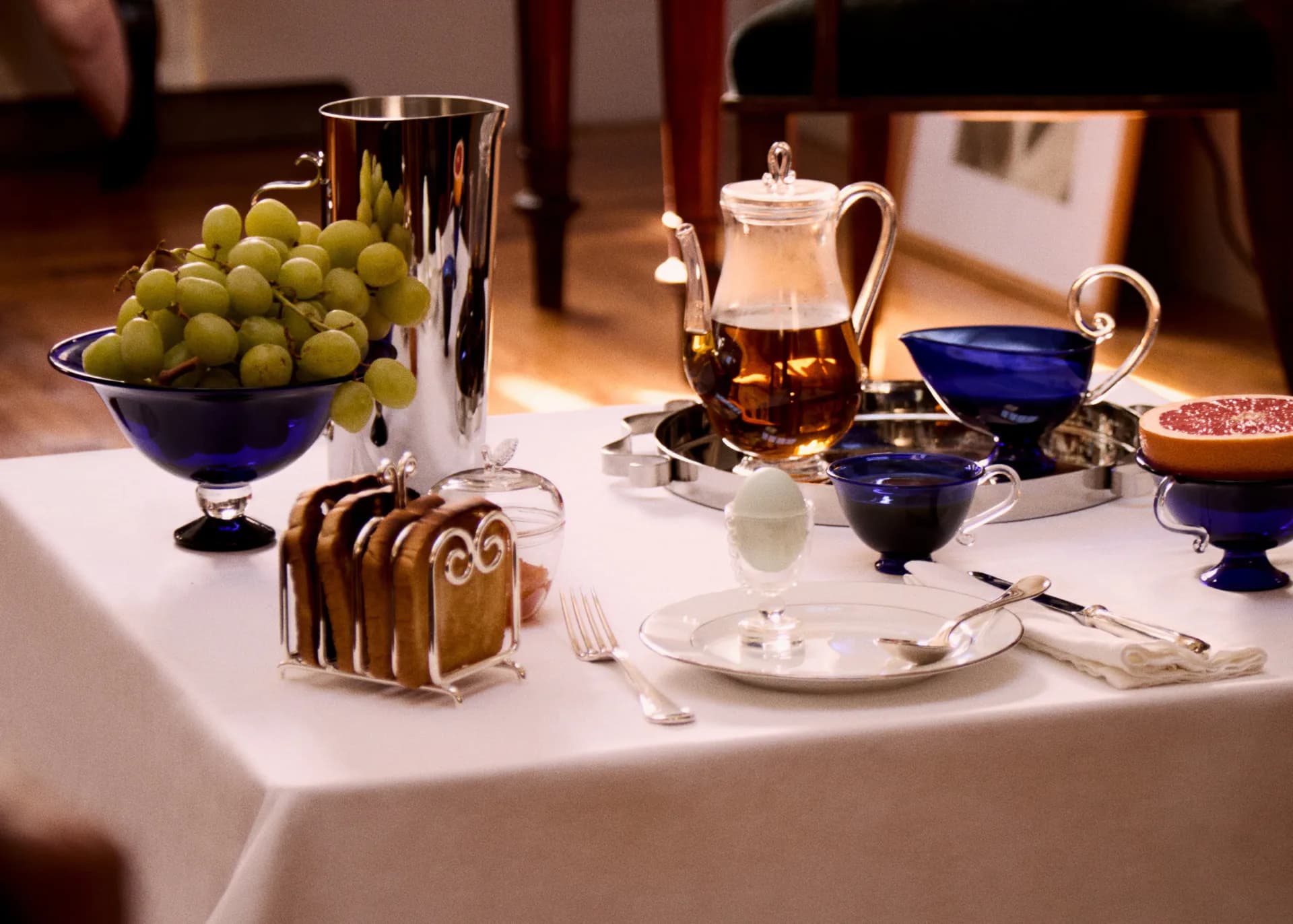 Elegant table setting with blue ceramic vessels, green grapes in a pedestal bowl, striped cake, and white linens, designed by Sophie Lou Jacobsen.
