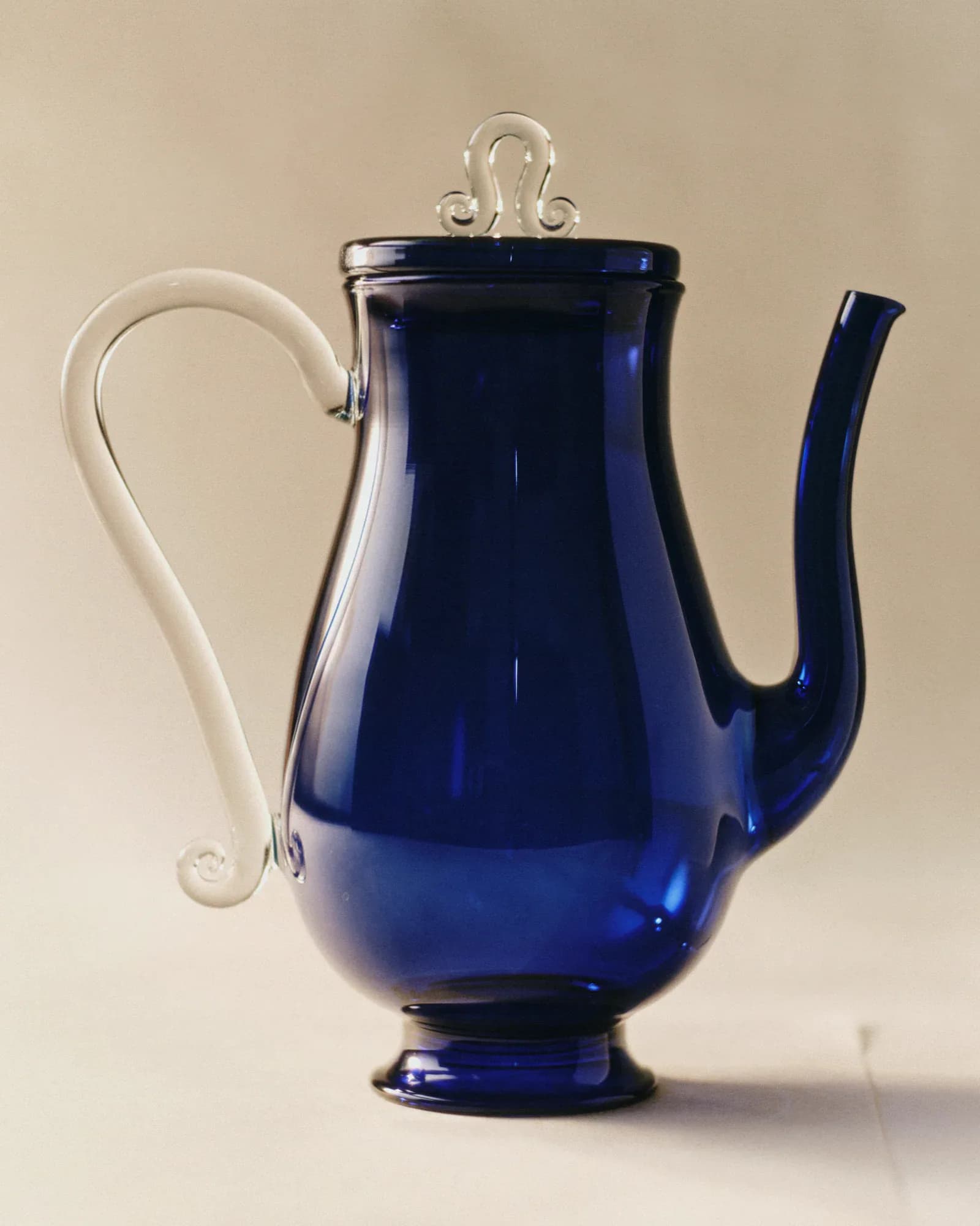 Cobalt blue glass teapot with clear glass handle and finial, displayed against a neutral beige background by Sophie Lou Jacobsen.