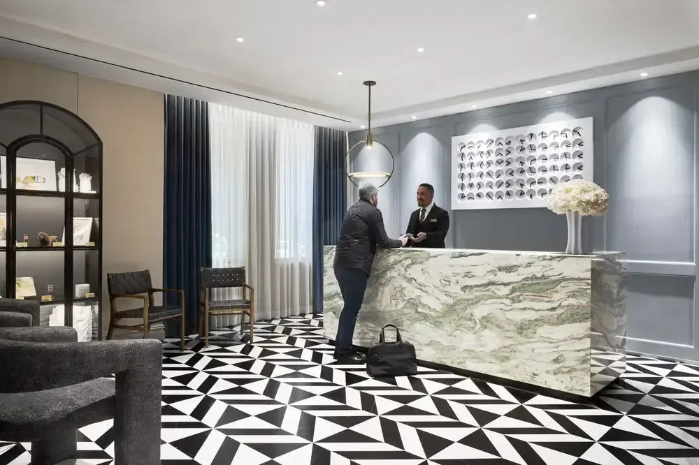 Modern hotel lobby with marble reception desk, geometric black-and-white patterned flooring, gray accent walls, and two figures at check-in, designed by Stacey Gilles.