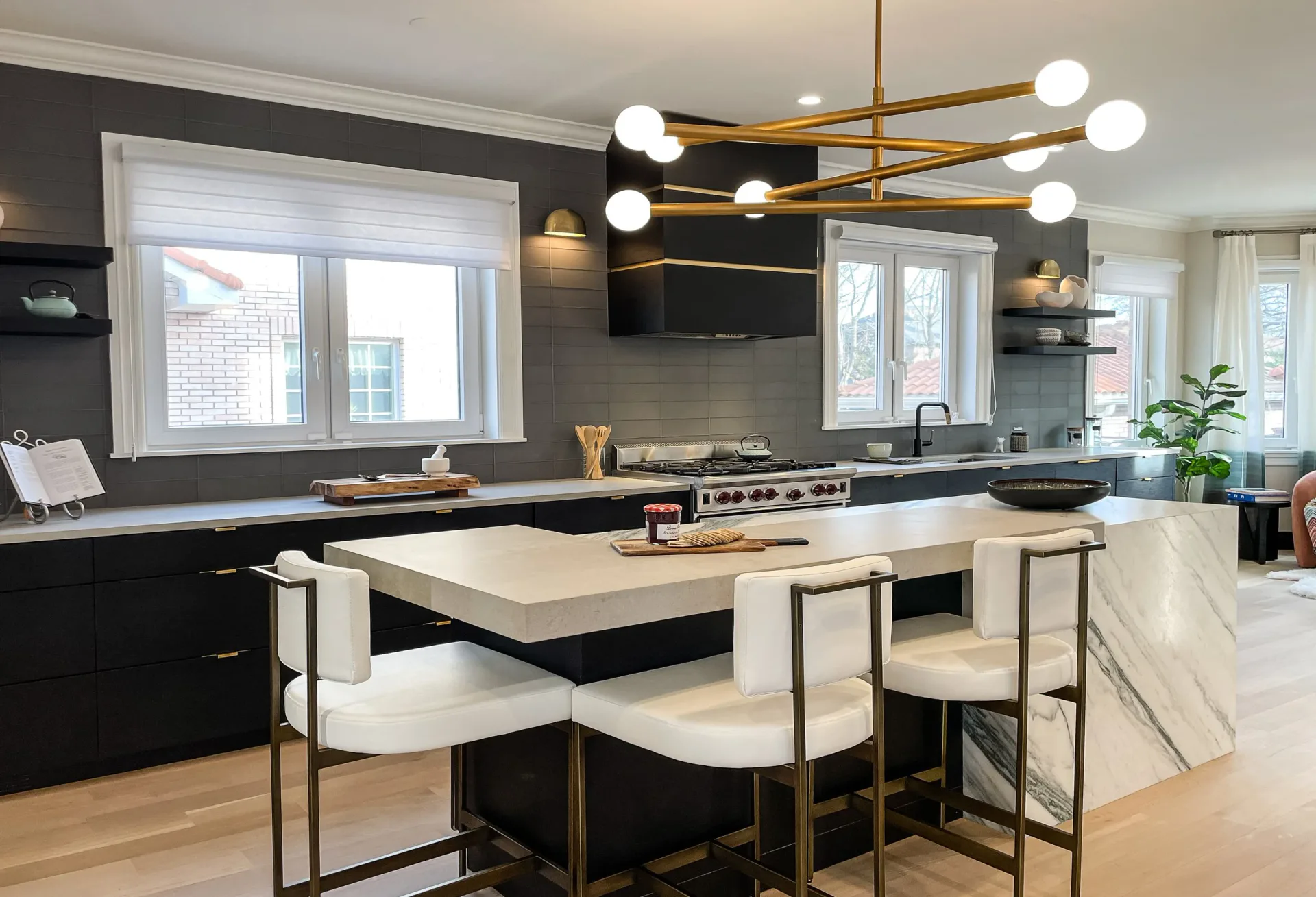 Modern kitchen with black cabinetry, white waterfall island, brass globe pendant lighting, and dark accent wall by Stavria Fourouli.