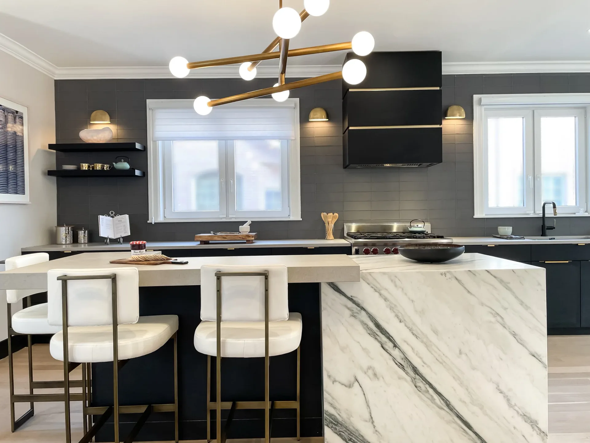 Modern kitchen with black cabinetry, marble waterfall island, brass branching chandelier, and cream upholstered bar seating by Stavria Fourouli.