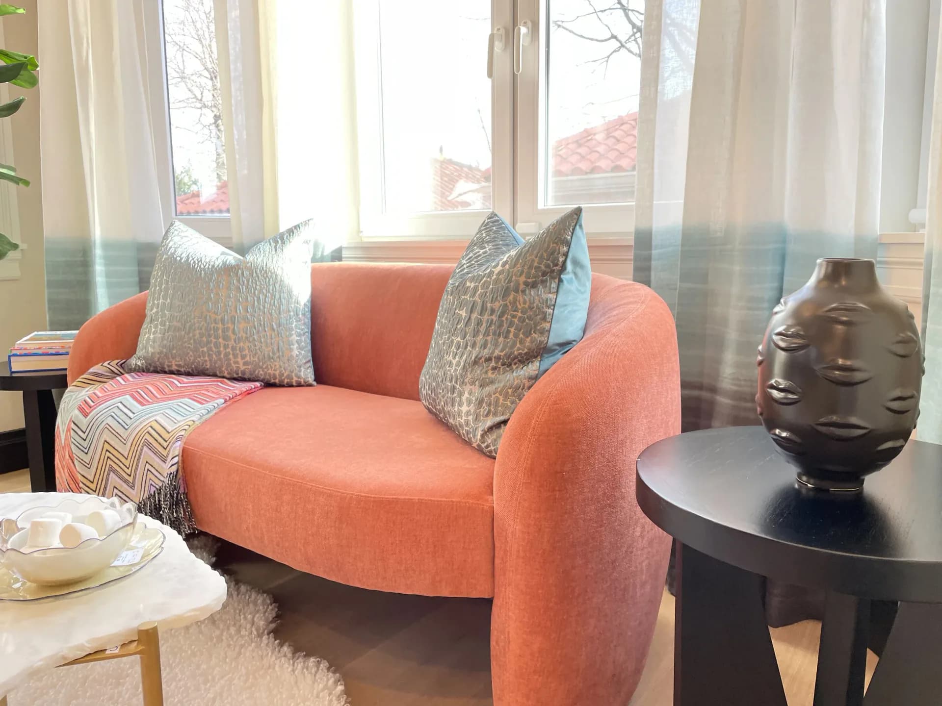 Bright contemporary living room featuring a sculptural orange curved armchair with gray textured pillows, black side table with bronze head sculpture, and sheer curtains framing large windows by Stavria Fourouli.