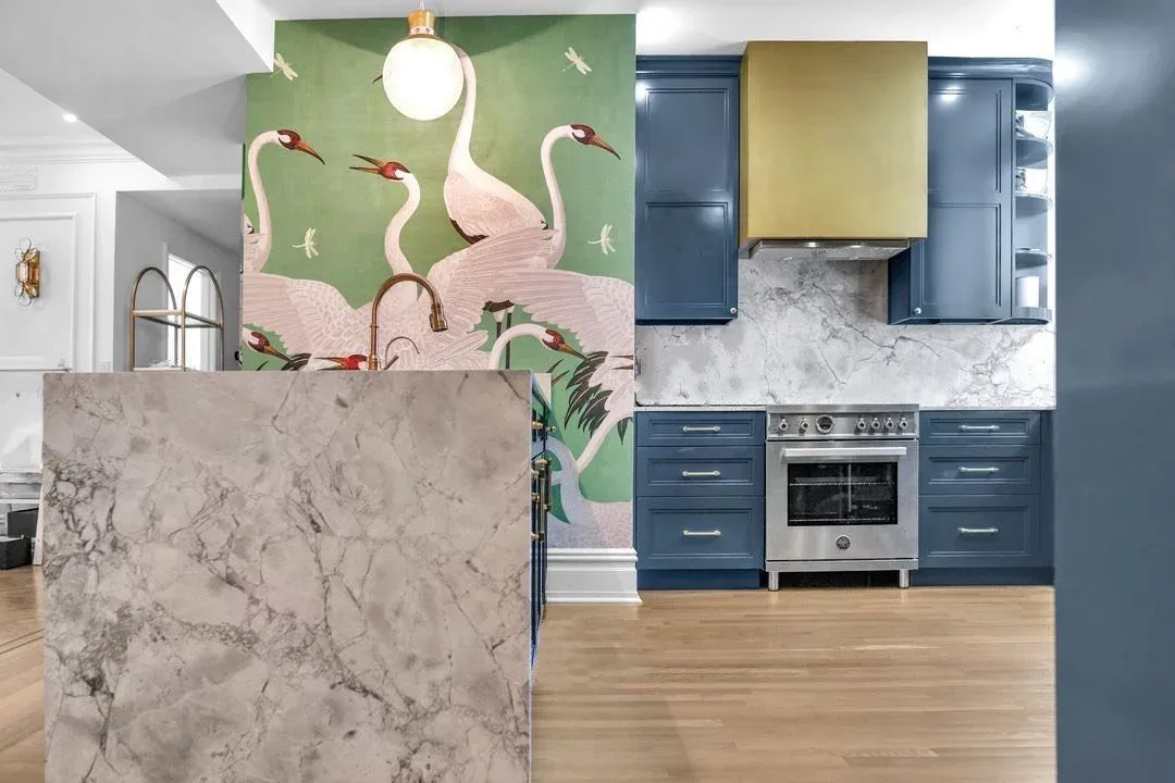 Modern kitchen with navy cabinetry, marble island, and brass hood, featuring a green accent wall with painted crane motifs by Stavria Fourouli.