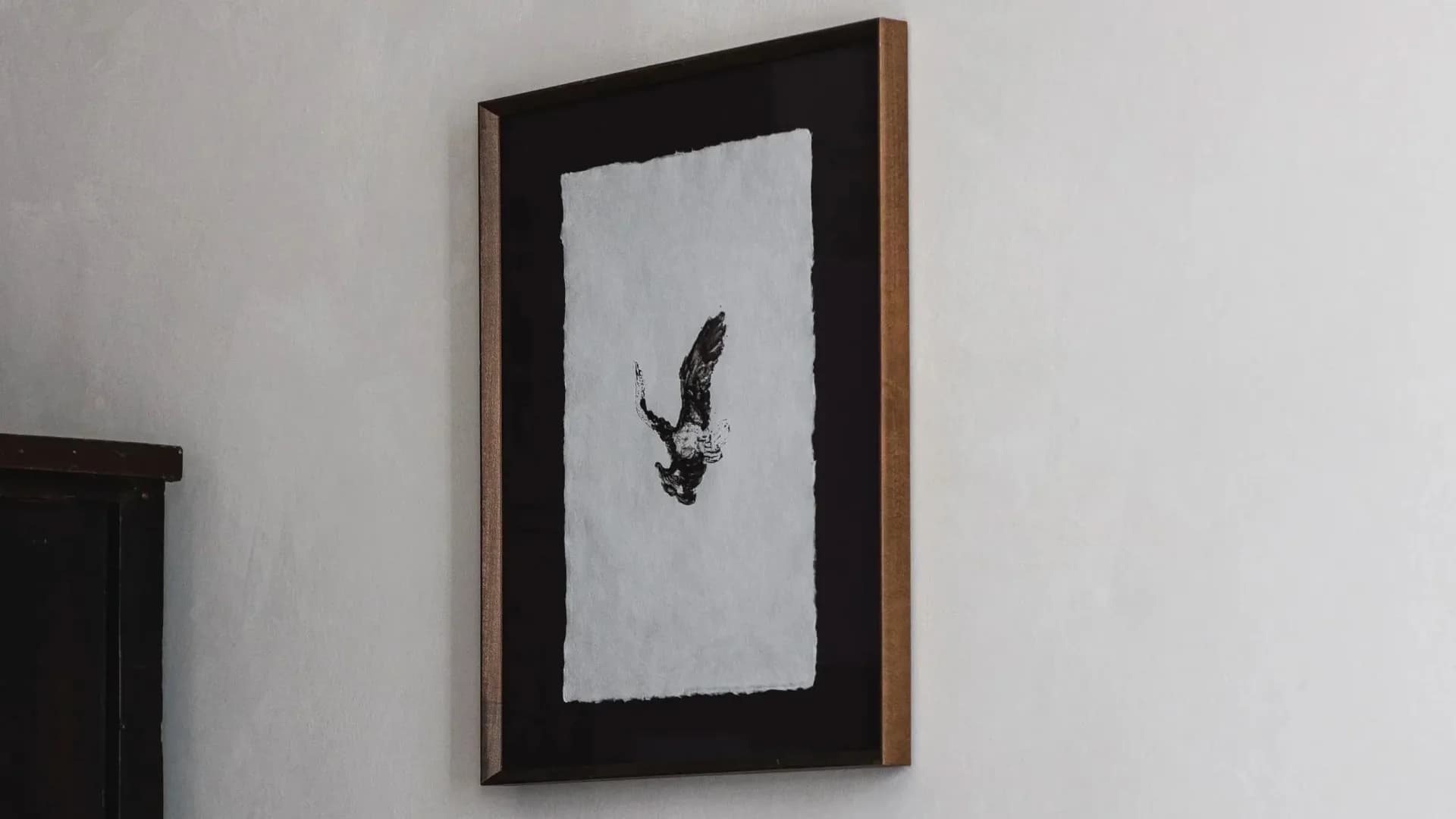 Framed artwork with black mat and wooden edge displaying a delicate feather sketch on white paper, installed on a light wall by Stefania Papadakis.