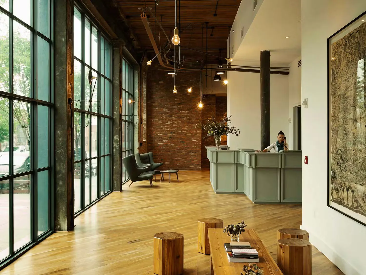 Modern commercial interior with exposed brick accent wall, polished wood flooring, green-framed glass doors, wooden ceiling, and minimalist furnishings by Stefanie Brechbuehler.