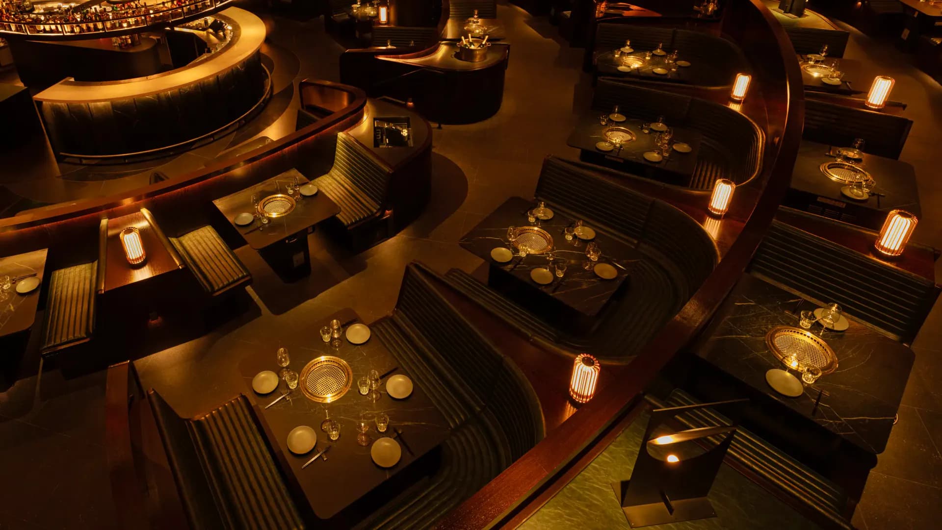 Intimate restaurant or bar interior with warm golden lighting, black tables with place settings, and glowing candle displays arranged throughout the space by Stephan Ritzer.