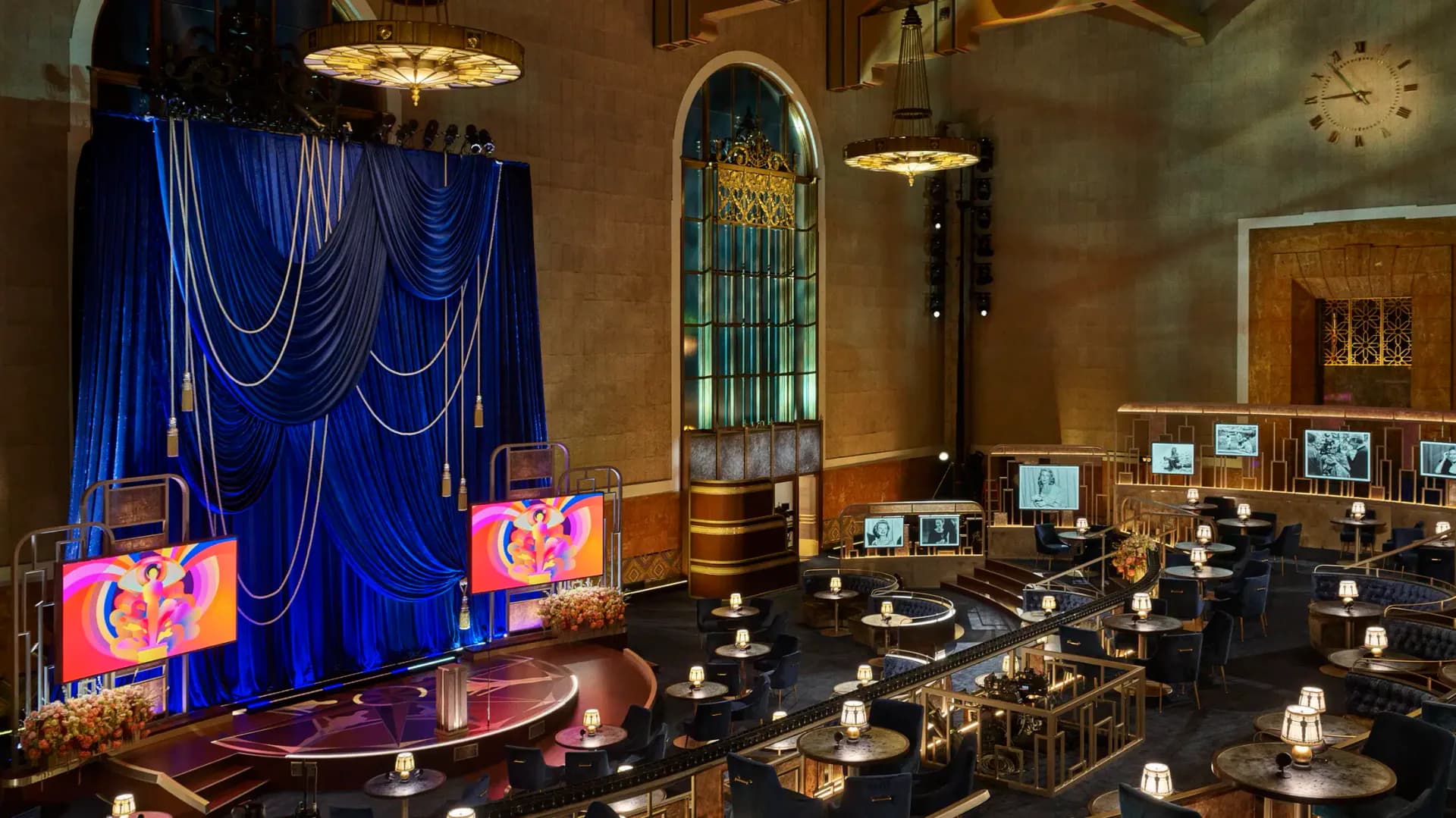 Elegant event space with dramatic blue draped stage, golden chandeliers, black round tables with candlelight, and art deco architectural details by Stephan Ritzer.