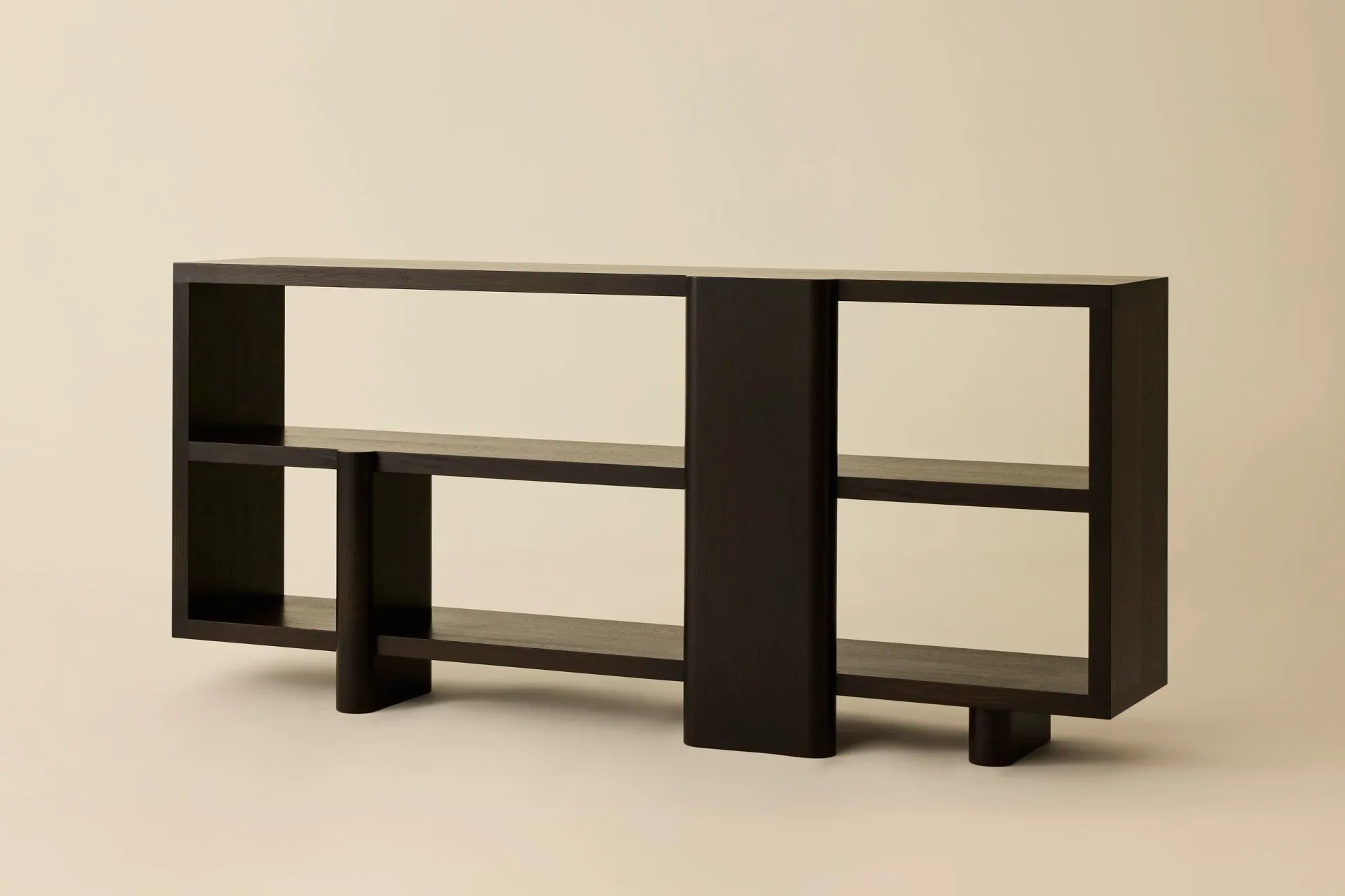 Dark wooden modular shelving unit with clean geometric lines and open compartments against a neutral beige background, a furniture design by Stephanie Beamer.