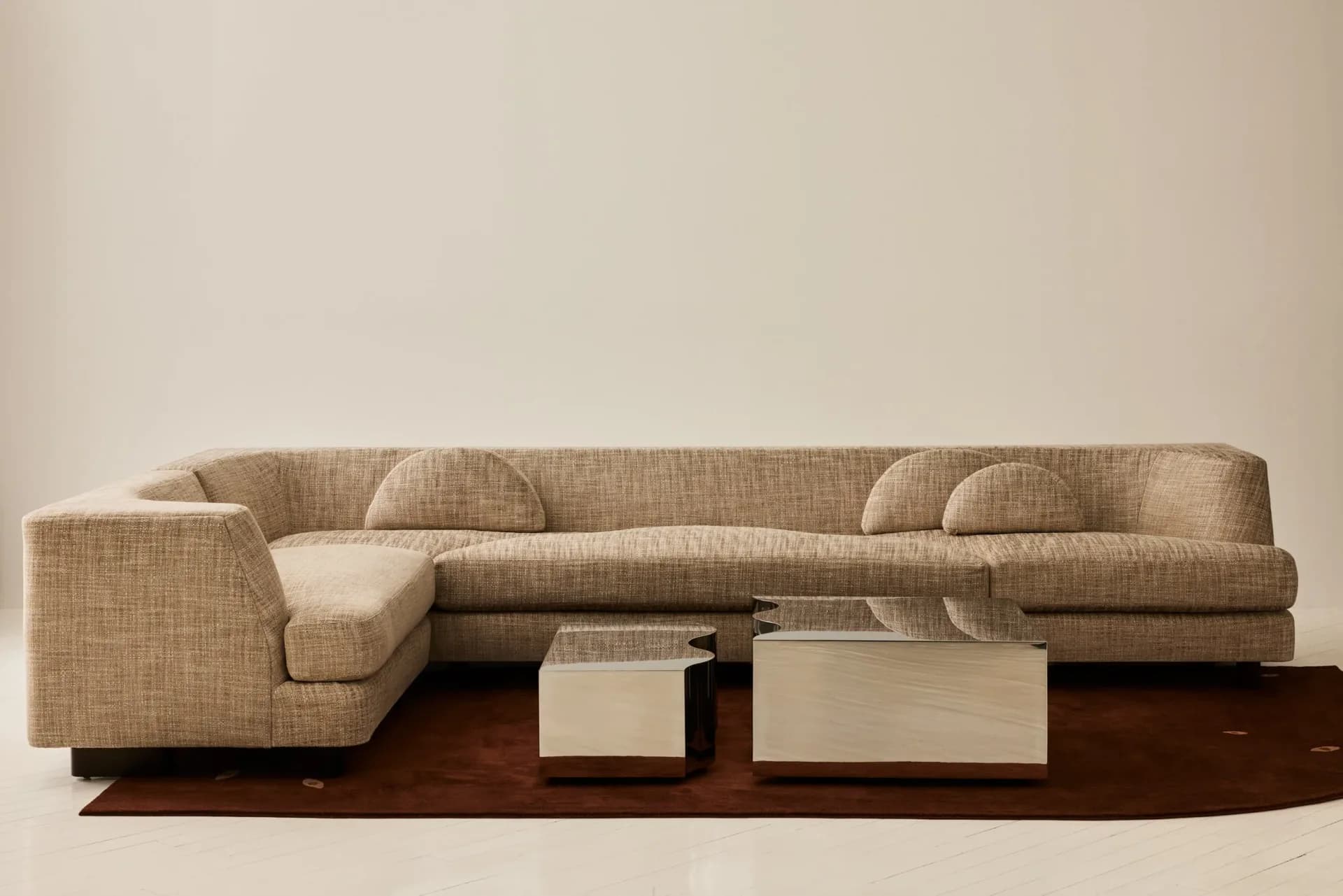 Beige upholstered modular sectional sofa with coordinating pillows and cream side tables on a dark wood platform, rendered in soft studio lighting by Stephanie Beamer.