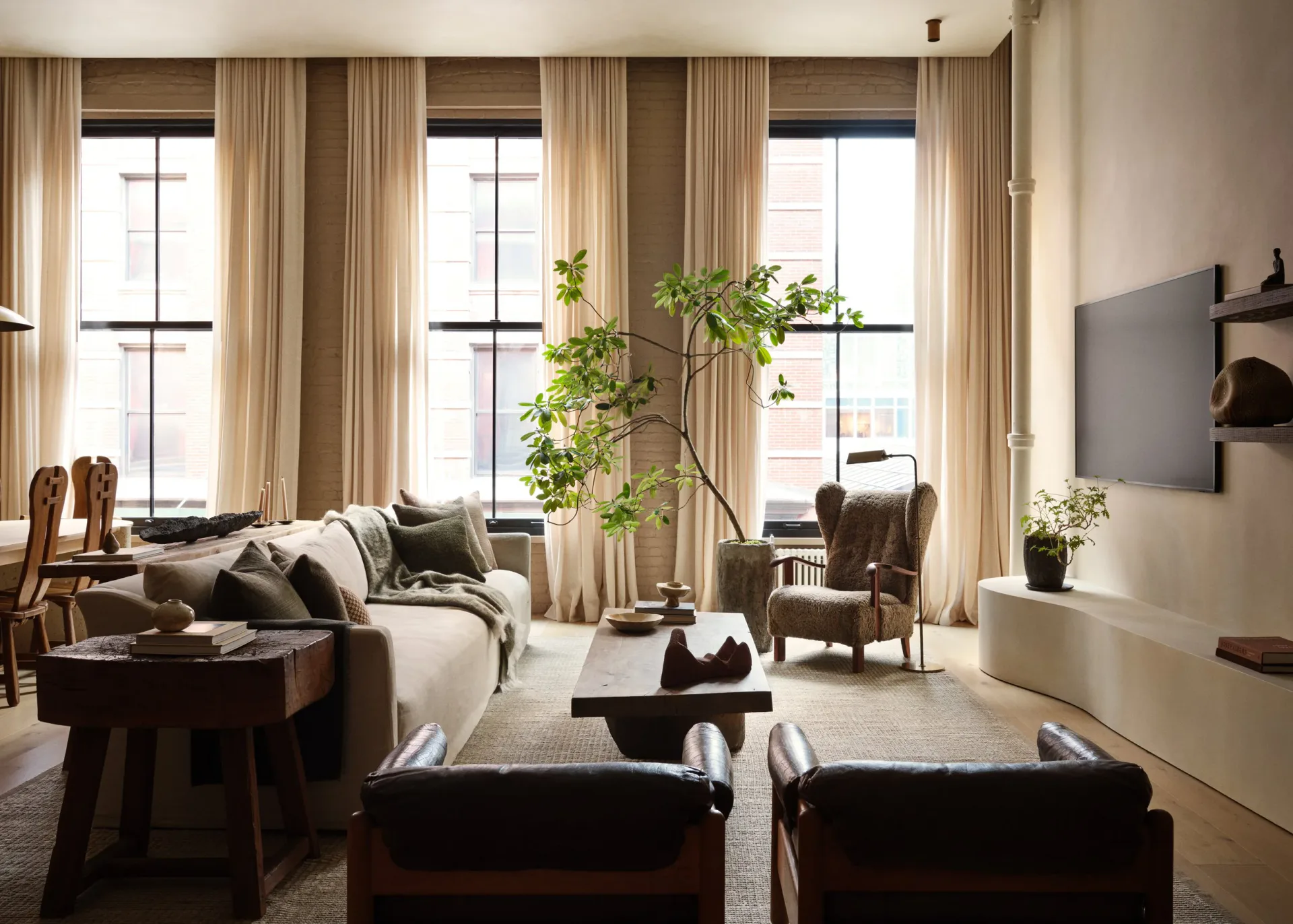 Spacious living room with high ceilings, tall windows with cream curtains, neutral palette, featuring a sofa, leather ottomans, green plant, and mounted TV by Stephanie Betesh.