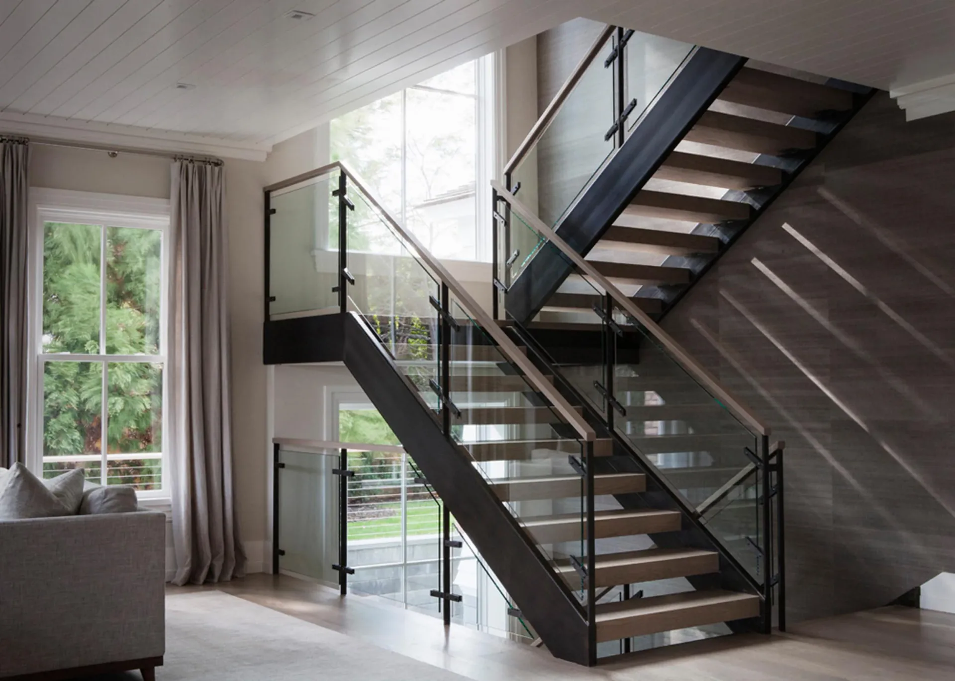 Modern multi-story staircase with black steel frame, glass railings, and wood treads in bright, minimalist interior by Stephanie DiTullio.
