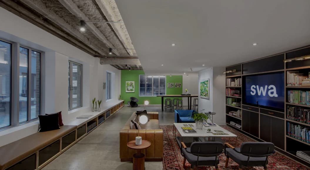 Modern office interior with exposed concrete ceiling, natural wood furniture, green accent wall, built-in shelving, and blue accent seating by Stephanie Gibson-Roberts.