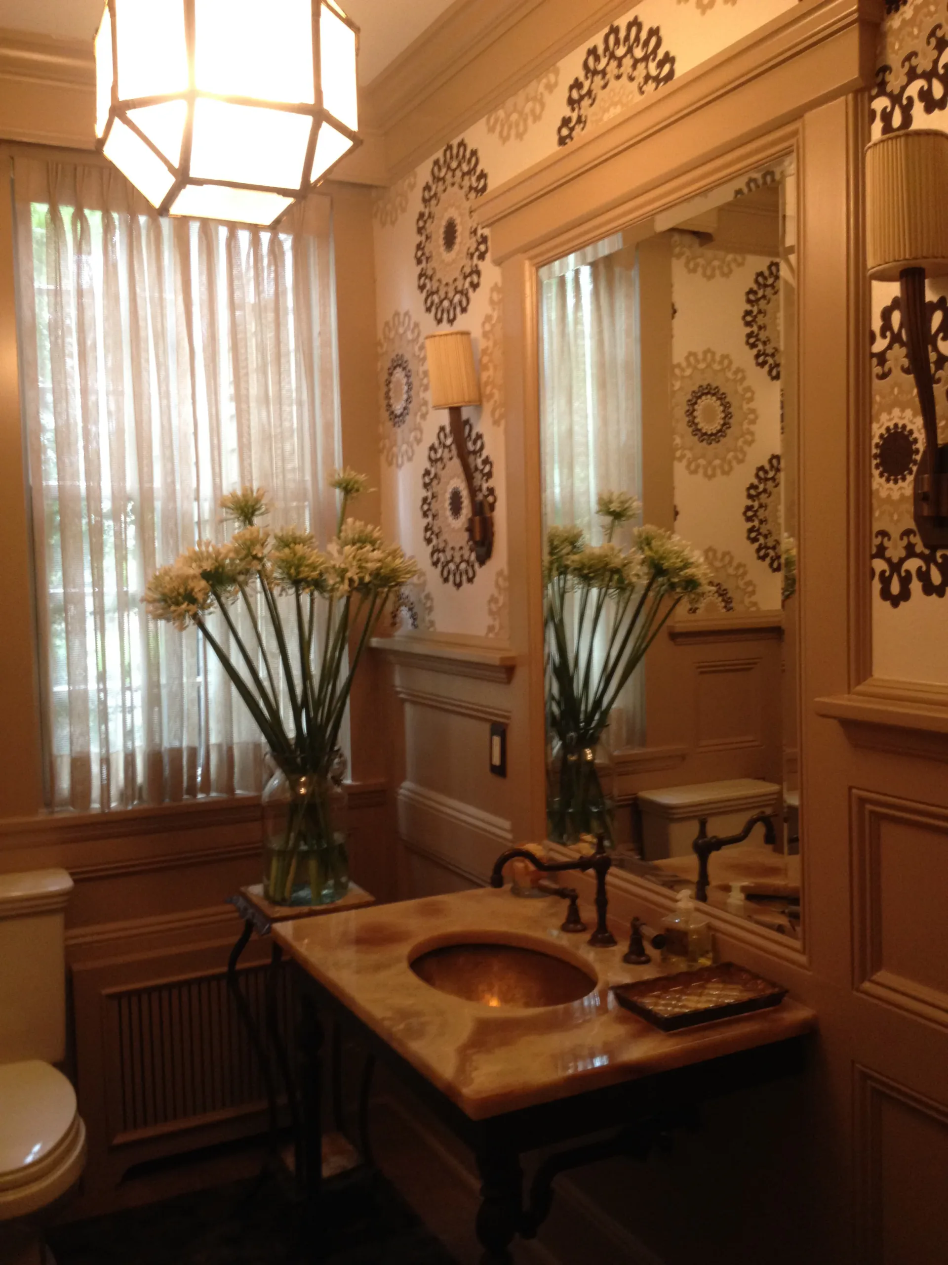 Elegant bathroom with warm wood vanity, decorative wall medallions, large mirror, brass fixtures, and floral arrangements by Stephanie Lichtenstein.