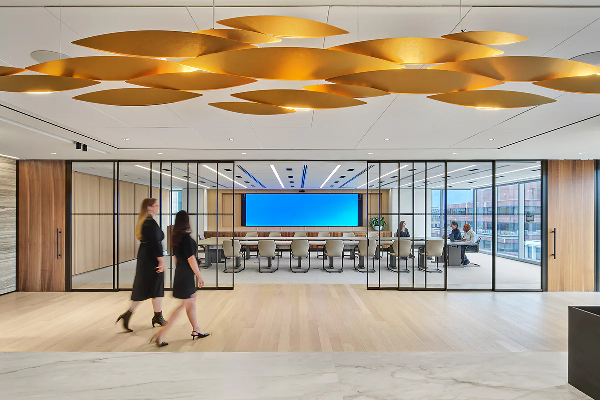 Modern office lobby with golden leaf-shaped ceiling fixtures, glass partition walls, and two figures walking toward a blue-screen meeting room by Stephen Bernstein.
