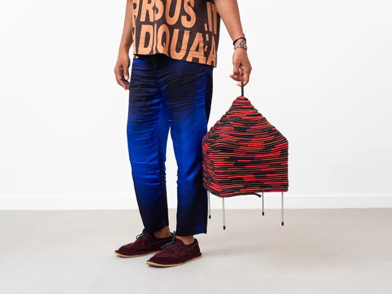 Model wearing orange graphic tee and blue patterned pants holds a red and black striped fabric chair design by Stephen Burks.