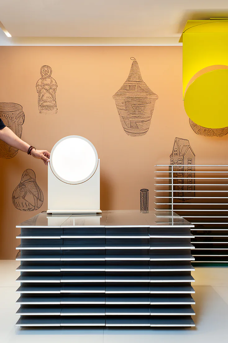 Warm peach wall with pen-drawn ethnographic vessel sketches, white circular mirror, stacked navy blue color sample cards, and yellow sculptural element by Stephen Burks.