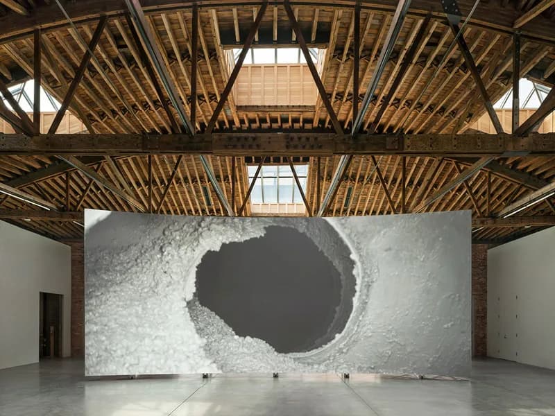 A minimalist gallery interior with exposed wooden ceiling beams and a dramatic white wall featuring a large dark circular void, designed by Stephen Cassell.