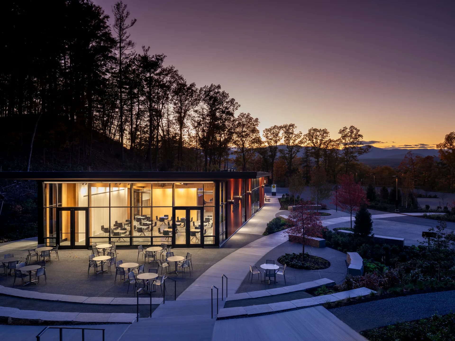 Modern illuminated pavilion with extensive glazing overlooks a twilight landscape with mountains, surrounded by contemporary hardscaping and trees by Stephen Cassell.