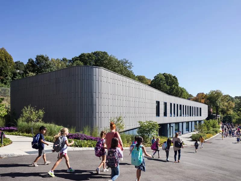 Modern institutional building with dark corrugated metal and light stone facade, featuring horizontal ribbon windows and a sloped roofline, surrounded by trees and landscaping with people in the plaza by Stephen Cassell