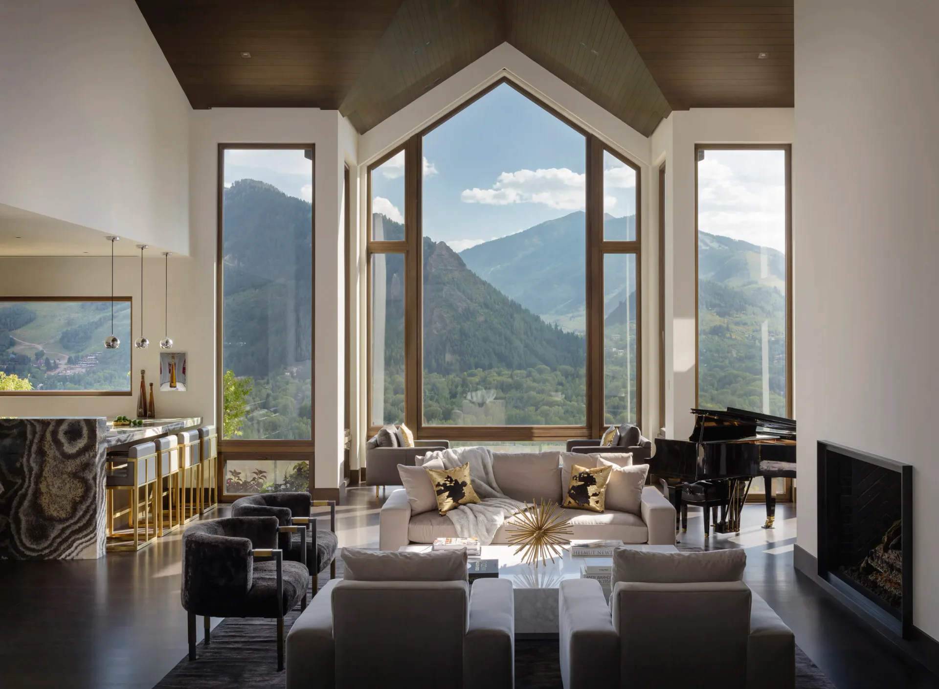 Modern luxury living room with soaring pitched-ceiling windows overlooking mountainous landscape, featuring taupe furnishings, grand piano, and dark flooring by Sterling McDavid.