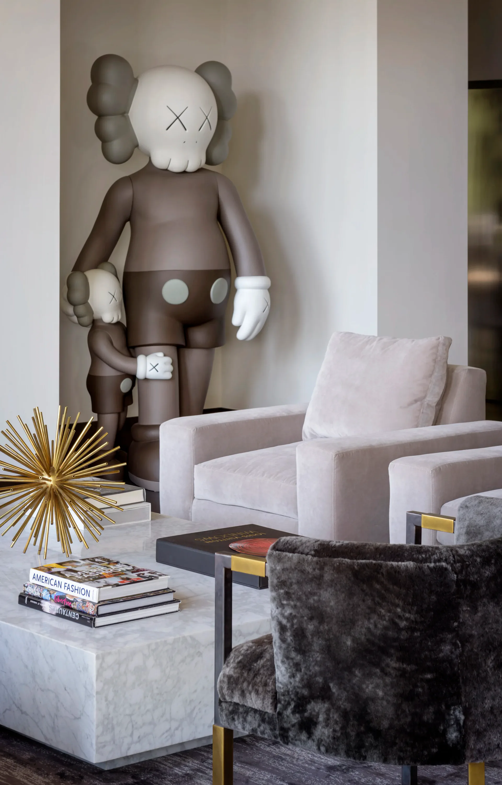 Modern living room interior featuring white upholstered armchairs, marble side table, and a striking oversized sculptural figure with X-marked face and gray tonal elements, designed by Sterling McDavid.