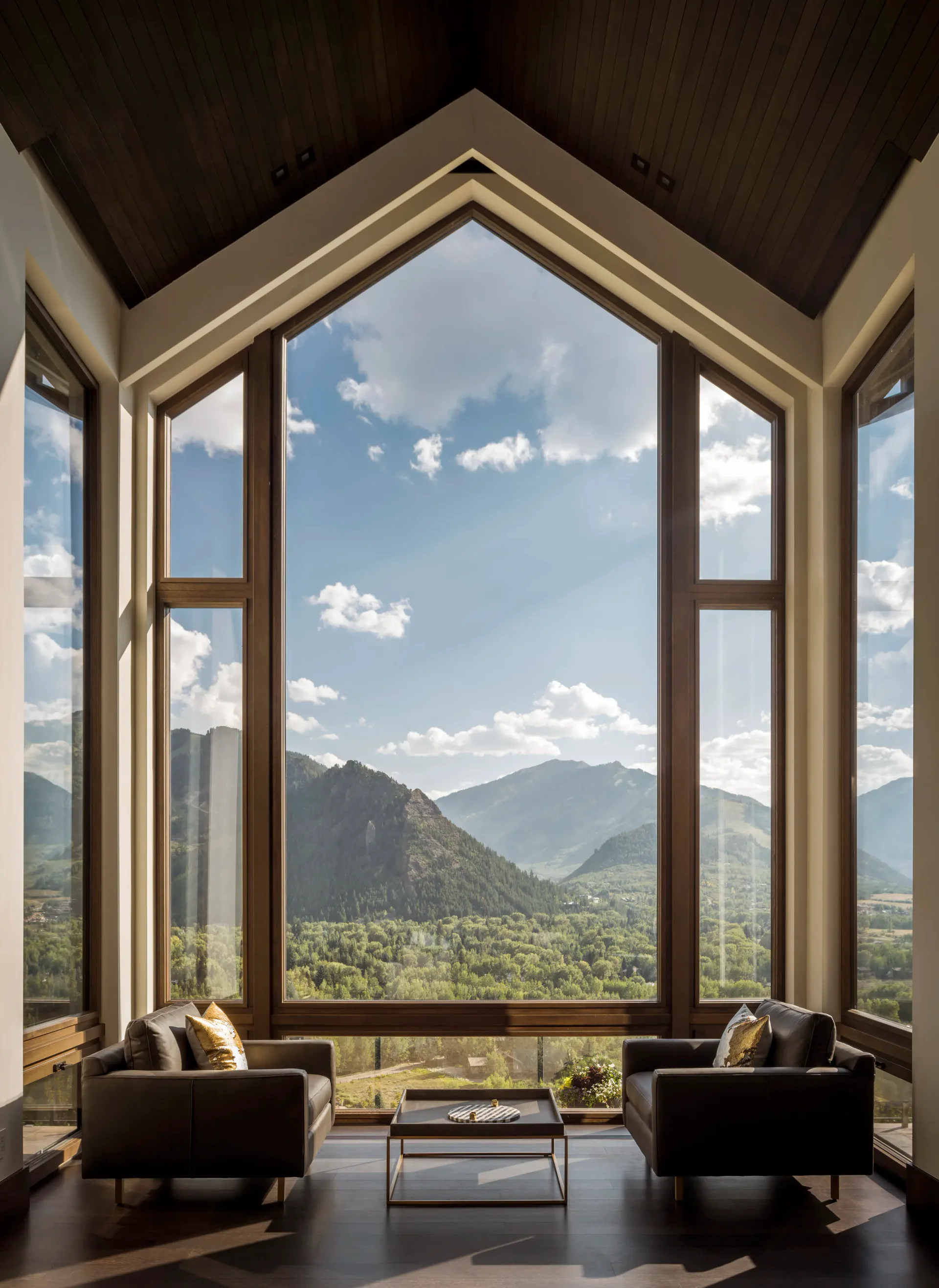 Modern living room with dramatic peaked window framing mountain valley views, dark furnishings, and warm wood ceiling by Sterling McDavid.