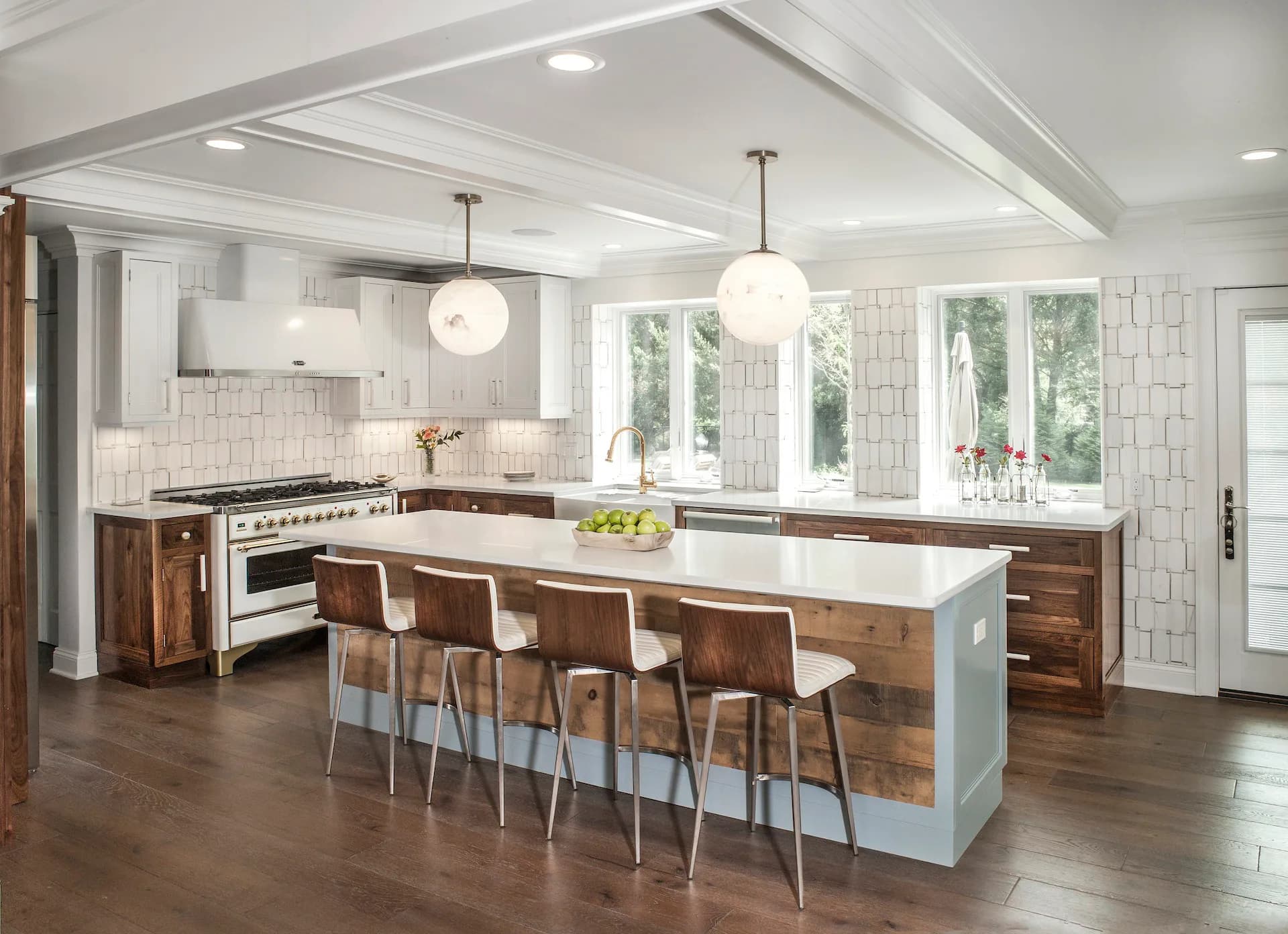 Modern kitchen with white subway tile backsplash, dark wood cabinetry, light blue island with seating, pendant lighting, and large windows overlooking trees by Steve Moran.
