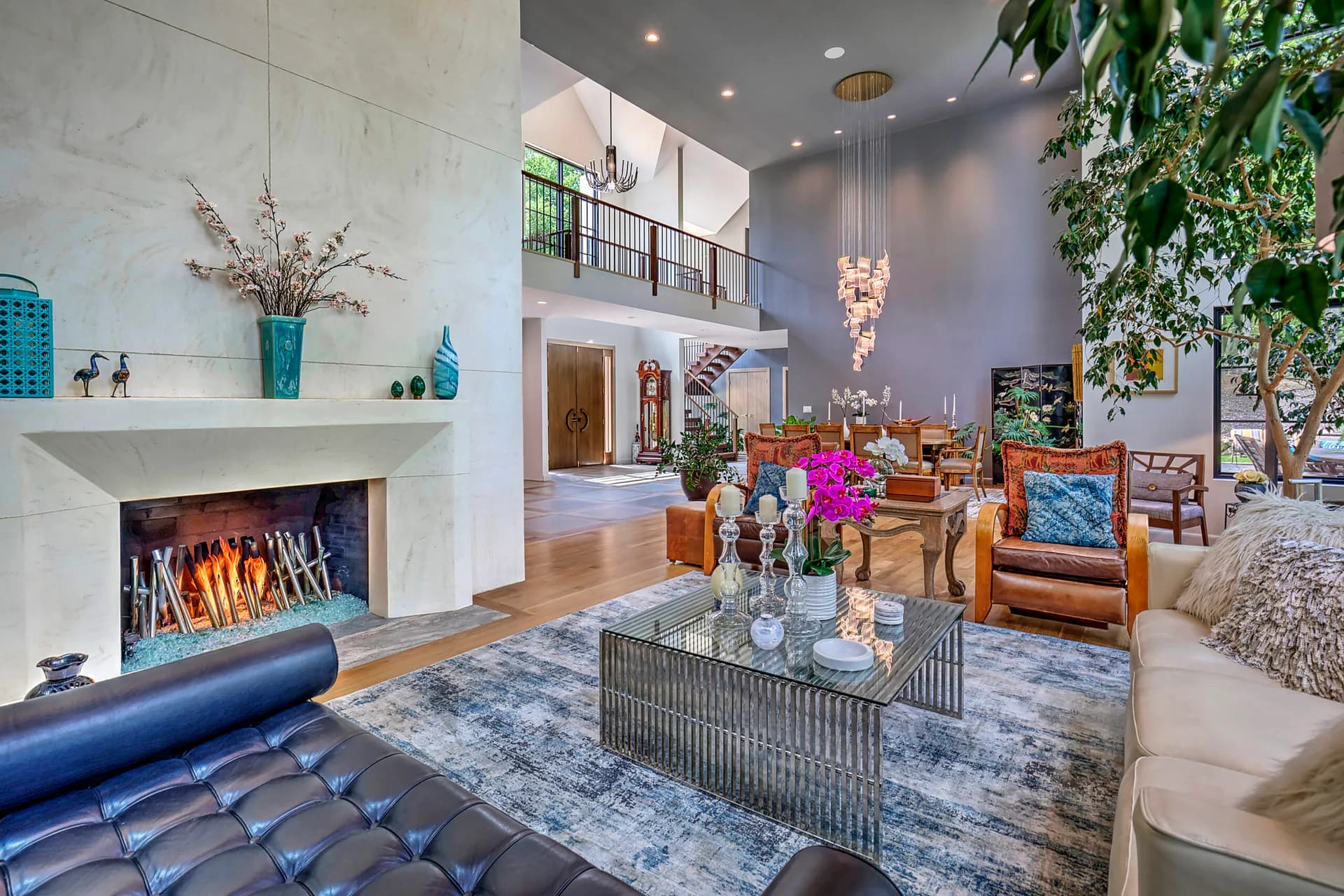 A double-height living room with fireplace, leather seating, geometric coffee table, vibrant floral arrangements, and lush greenery throughout, designed by Steve Moran.