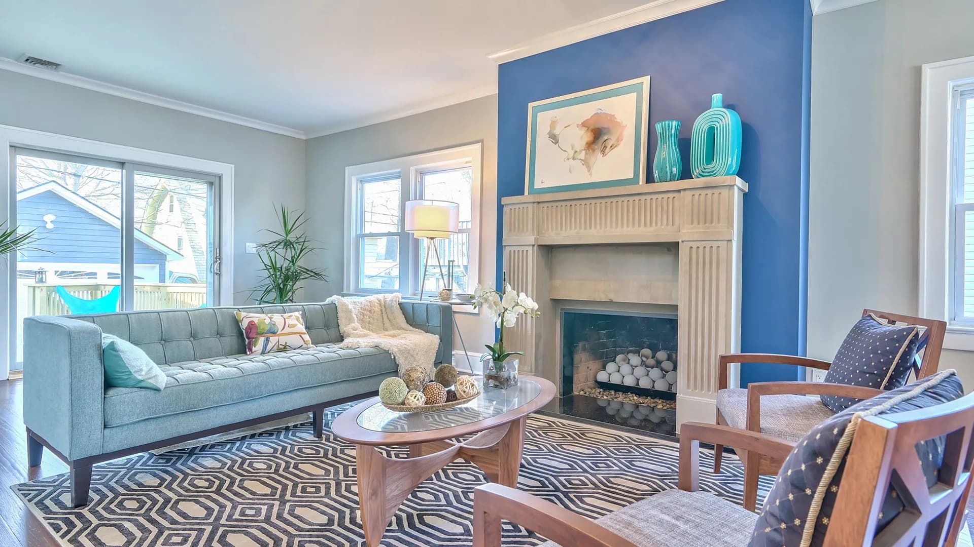 Bright living room with blue accent wall, fireplace, gray sofa, geometric patterned area rug, and mixed wood furniture by Steve Moran.