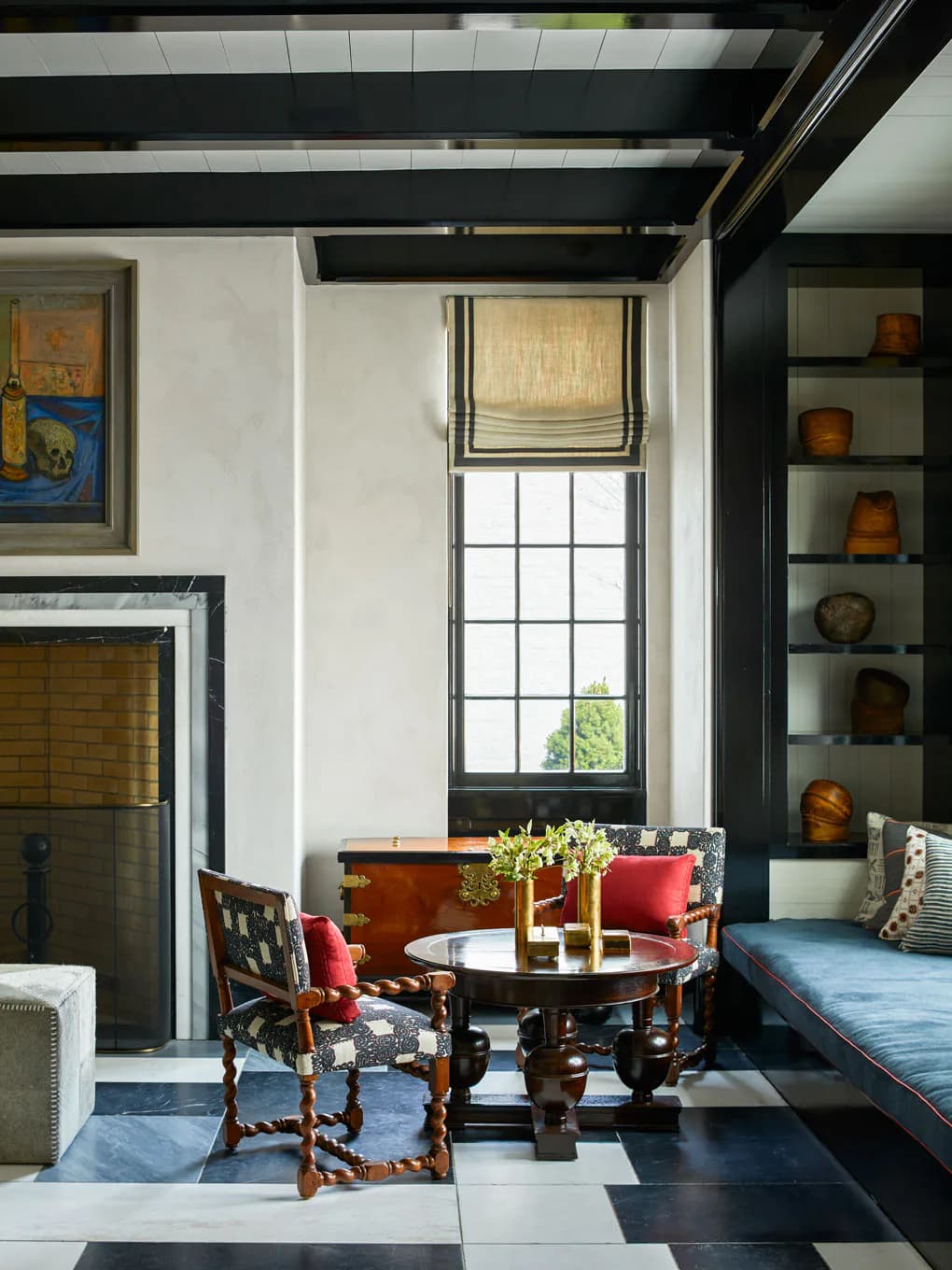 Contemporary interior with black exposed beams, white walls, checkered floor, round table with red upholstered chairs, built-in shelving displaying ceramics, and gridded window with Roman shade by Steven Gambrel.