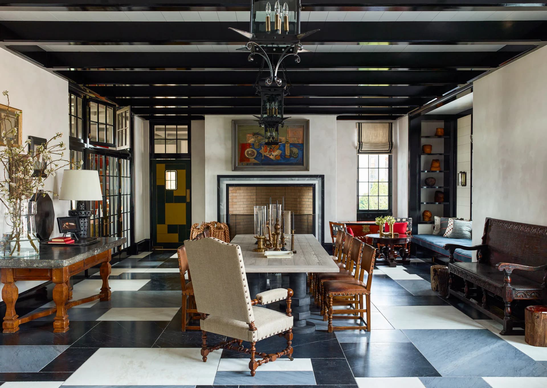 A sophisticated living and dining space features black exposed ceiling beams, white walls, a geometric black-and-white floor, period furniture, and a fireplace, designed by Steven Gambrel.