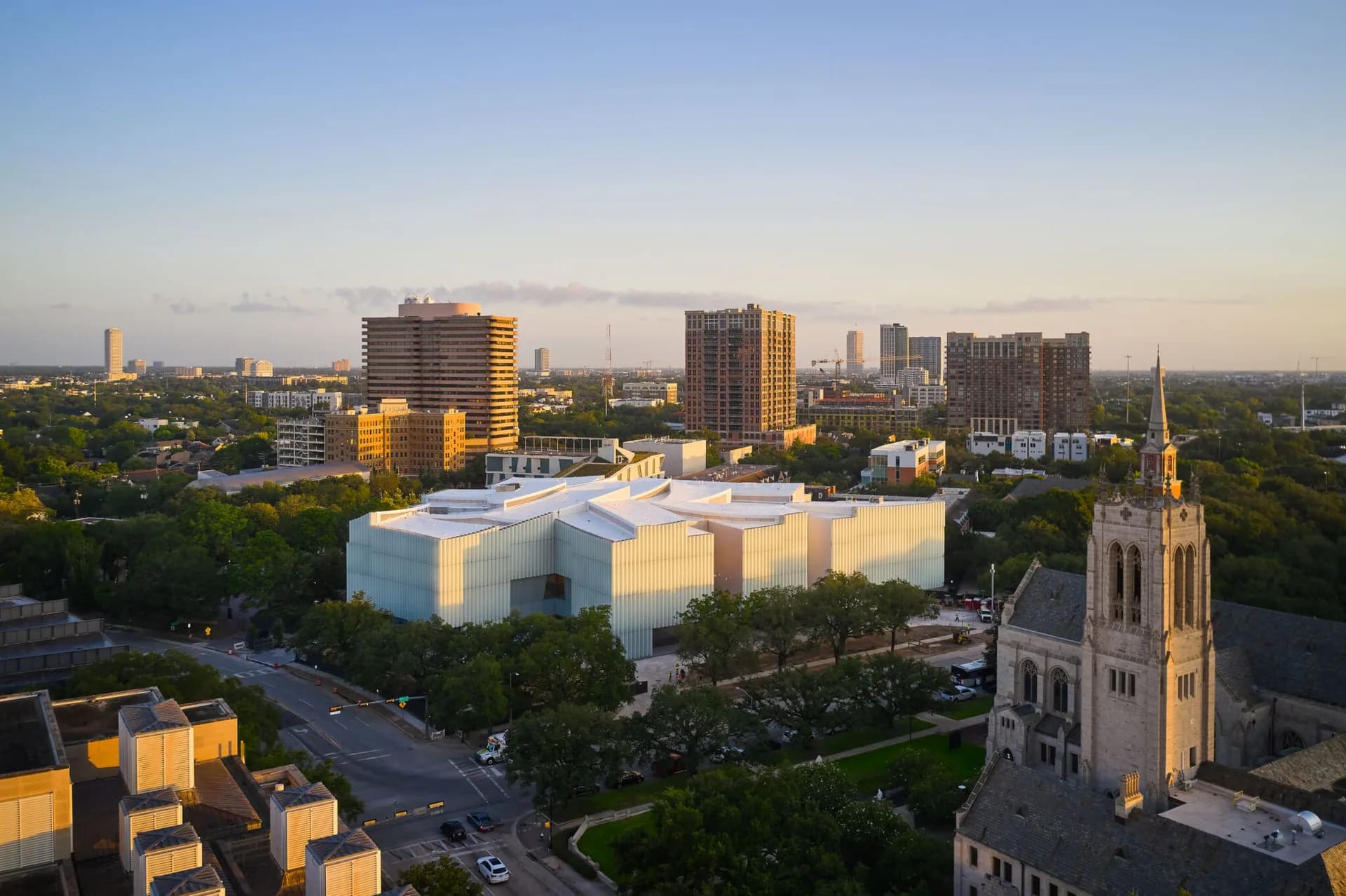 Aerial view of a modernist white cubic museum building with geometric forms surrounded by green landscape and historic church spire, photographed at sunset by Steven Holl.
