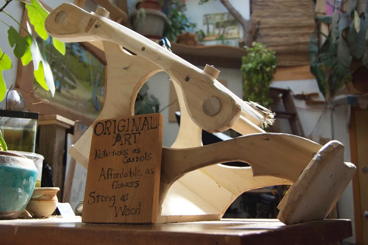 Wooden birdhouse with cardboard signage displaying design details, surrounded by potted plants and natural materials in a bright studio setting by Stewart Hoyt.