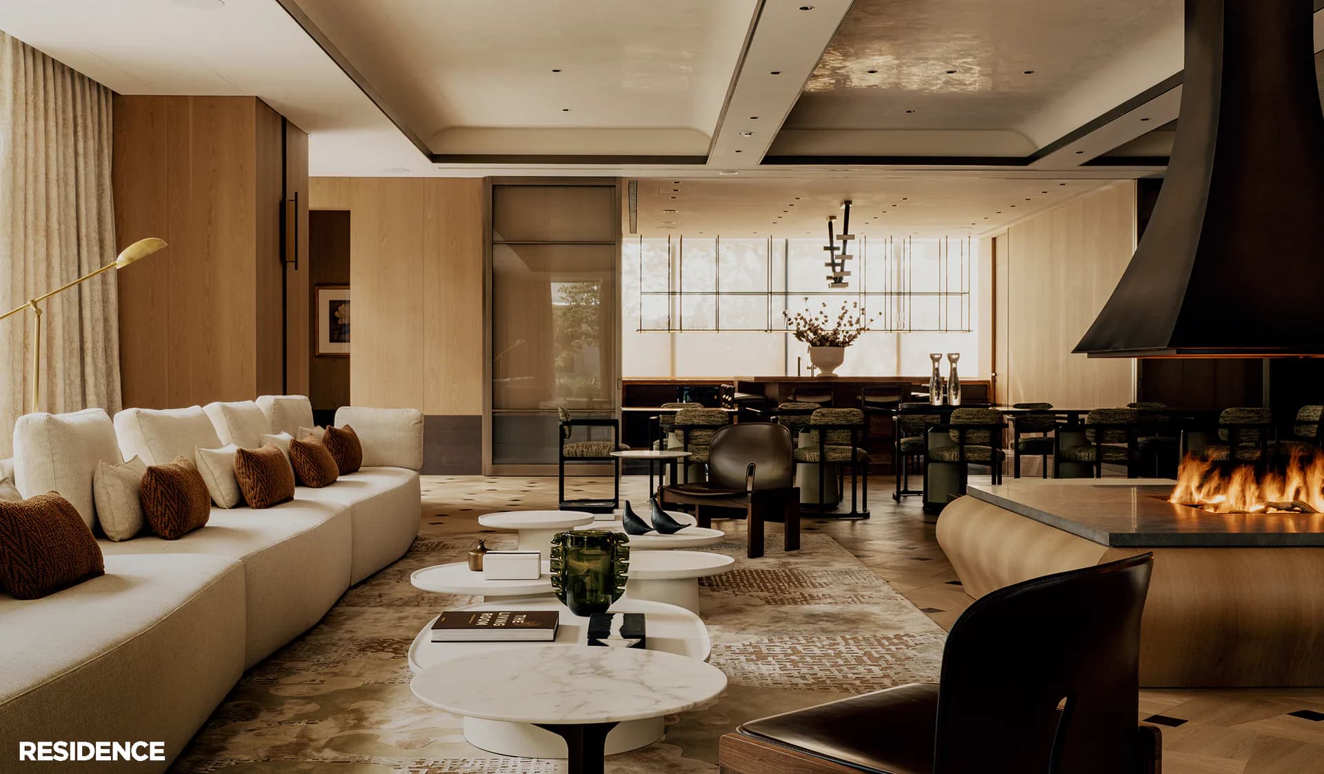 Luxurious open-plan residence with curved cream sofa, linear fireplace, dark dining table, warm wood paneling, and floor-to-ceiling windows overlooking cityscape by Su Ergeneli.